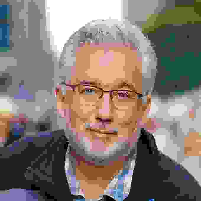 A portrait of Jack Reznicki, who is looking into the camera with a smile. Jack has silver hair, and he is wearing a pair of glasses with a black, thin frame. This photo of Jack is taken on a busy sidewalk.