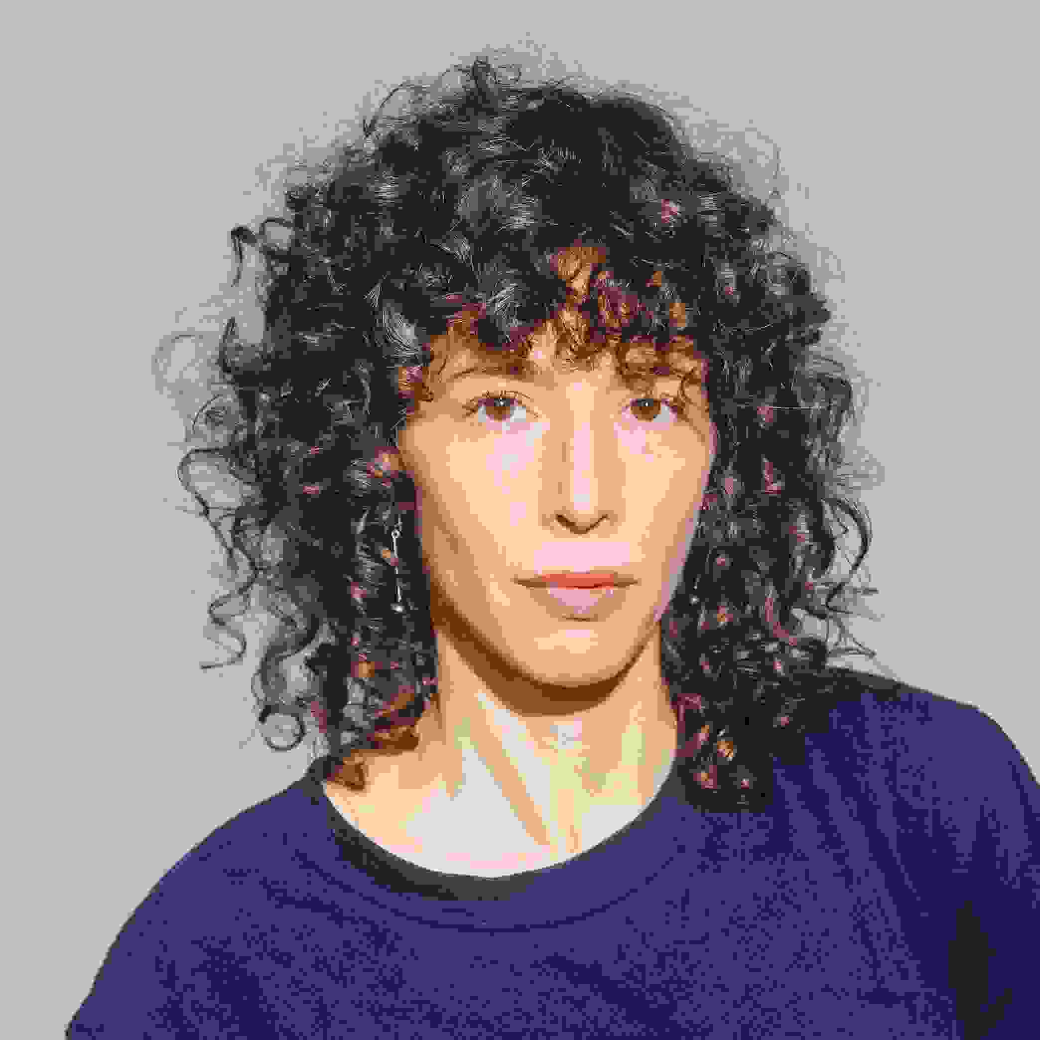 Image of a woman looking into the camera. The woman has curly brown hair and is wearing a blue shirt.