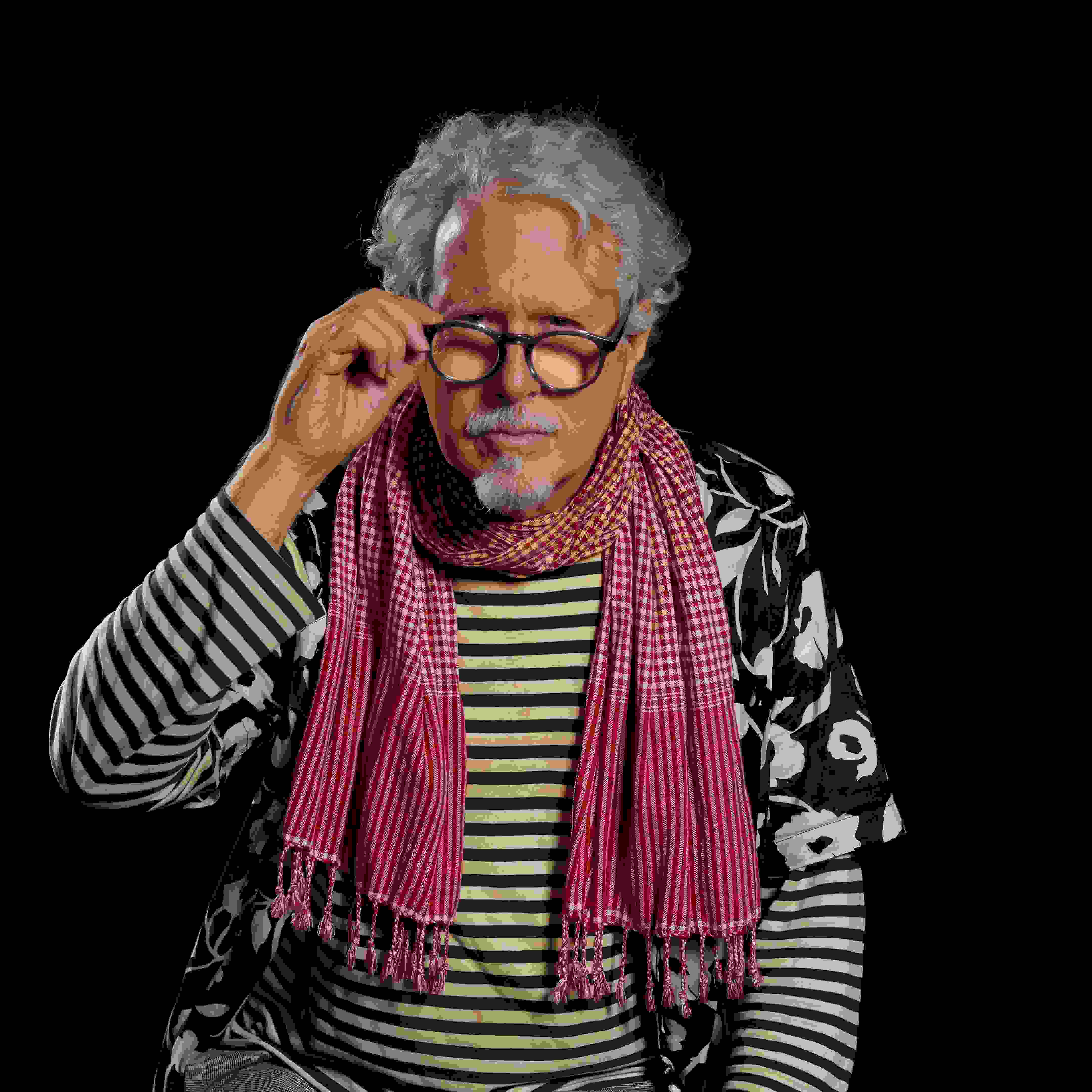 image of a man in front of a black background looking into the camera. the man is wearing a black and white striped shirt with a red gingham scarf and a black and white flowered hawaiian shirt. The man has white curly hair and wearing glasses.