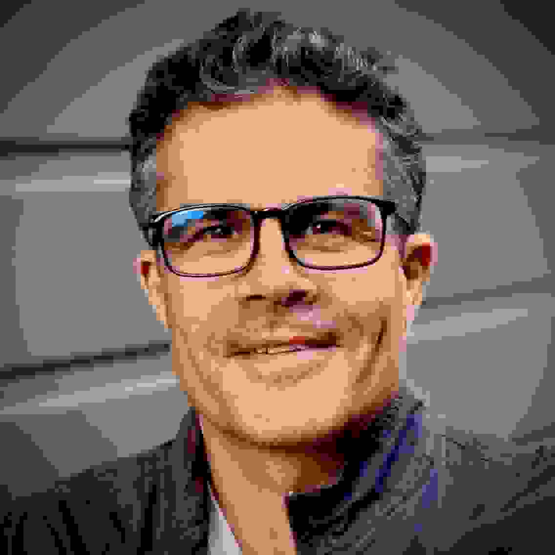 Color photo of Mark Tribe showing head and shoulders. He is wearing glasses, smiling, and shown against a gray background.