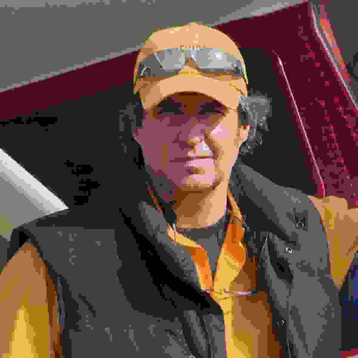 Image of a man looking at the camera. The man is wearing an orange shirt, a black puffy vest, an orange hat with sunglasses on top.