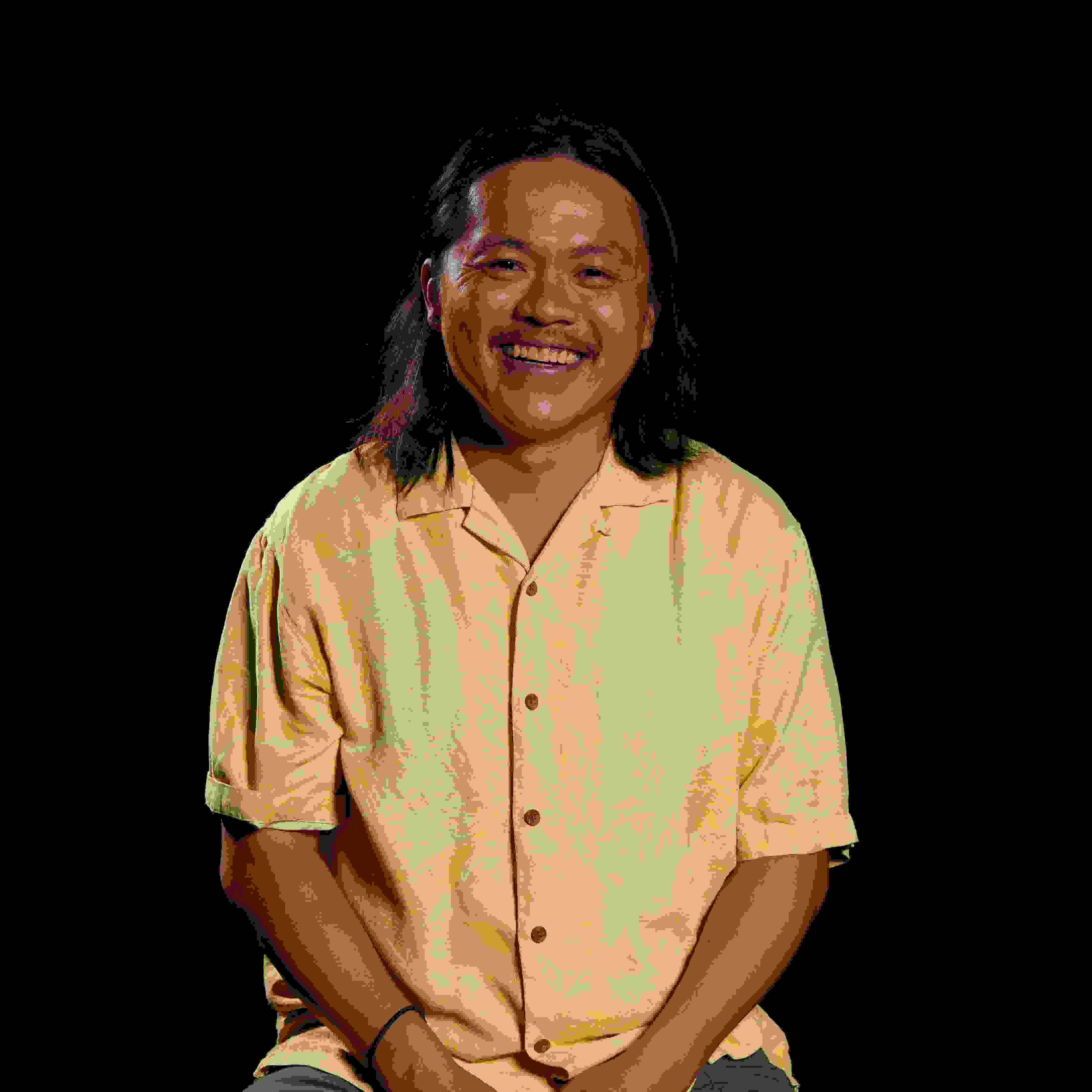 Image of a man smiling at the camera in front of a black background wearing a yellow shirt. The mean has shoulder length brown hair.