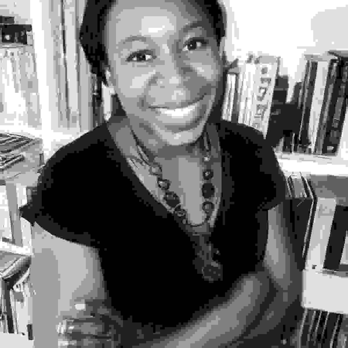 Black and white image of a woman smiling at the camera. The woman has her arms crossed, standing in front of a bookshelf wearing a black shirt and necklace.