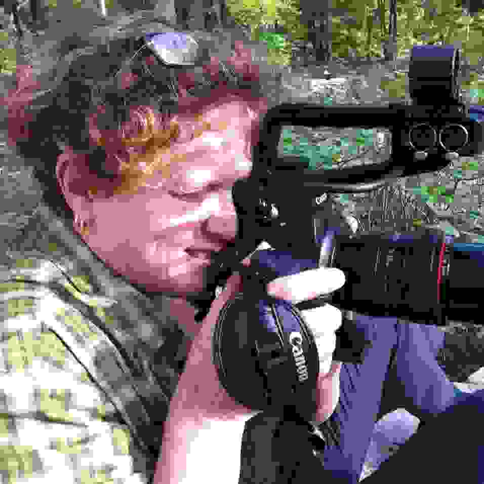 Image of a woman looking through a Canon video camera, shooting video. The woman is sitting on the grass, with curly red/brown hair, glasses on her head, and wearing a plaid shirt.