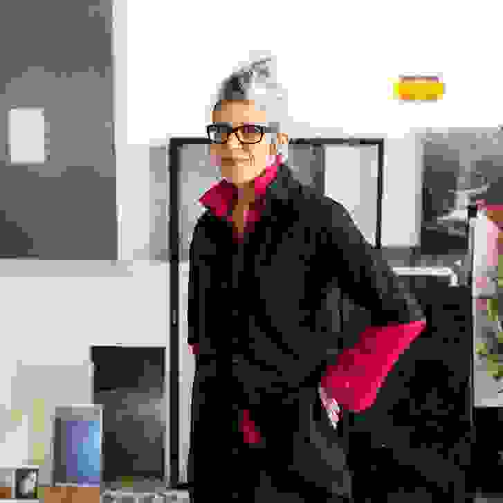 Image of a person standing in an art studio looking at the camera. The person is wearing a black button up with a pink undershirt and glasses.