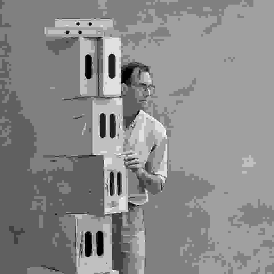 Black and white image of man standing behind a tall stack of studio "apple boxes" looking away. The man is wearing glasses.