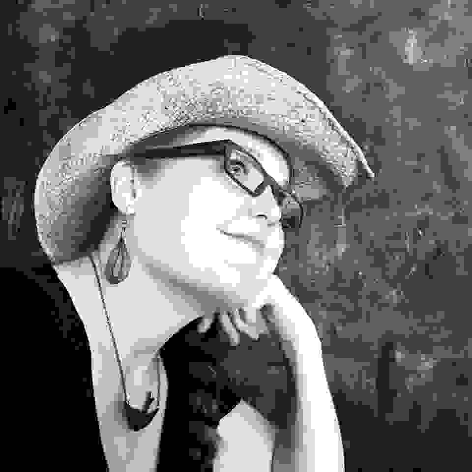 A back-and-white profile photo of Therese Gietler, who is looking to the top right corner of the frame. She wears a straw cowboy hat, a pair of glasses, a laced earing, and a necklace with a bird pendant.