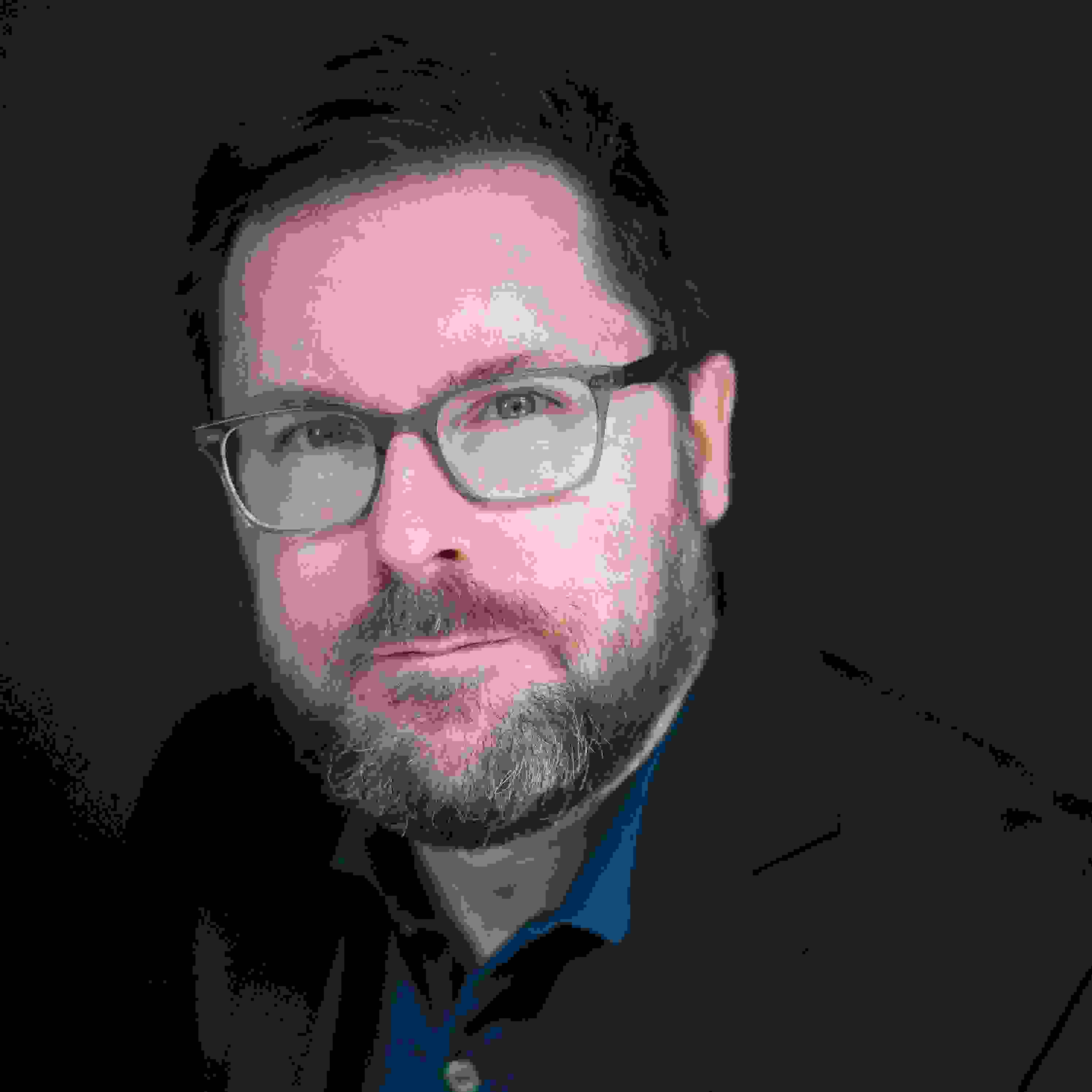 Portrait of Tom Ashe. Tom is wearing glasses and a blue jacket over a blue polo shirt.