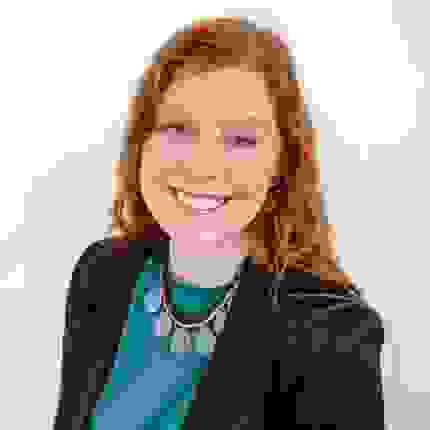A headshot of a woman with red hair, smiling, wearing a black blazer over a teal shirt with a chunky blue necklace, against a white background