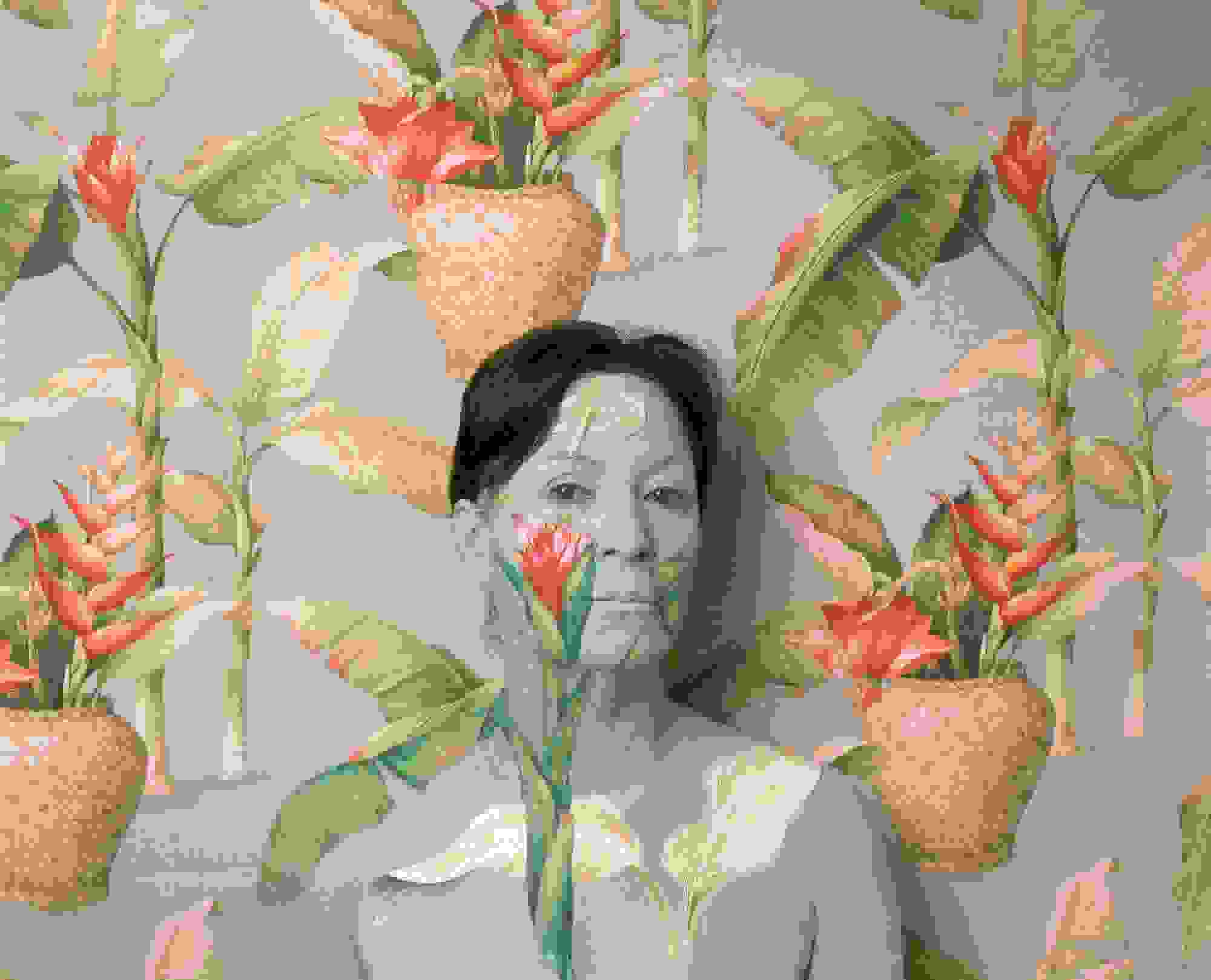 A photograph of a woman from the chest up standing in front of an ornately-wallpapered wall. The wallpaper is pale blue, and there are green plants with orange flowers sticking out of woven pots. The woman is painted with the same pattern to blend into the wallpaper, with only her brown hair standing out.