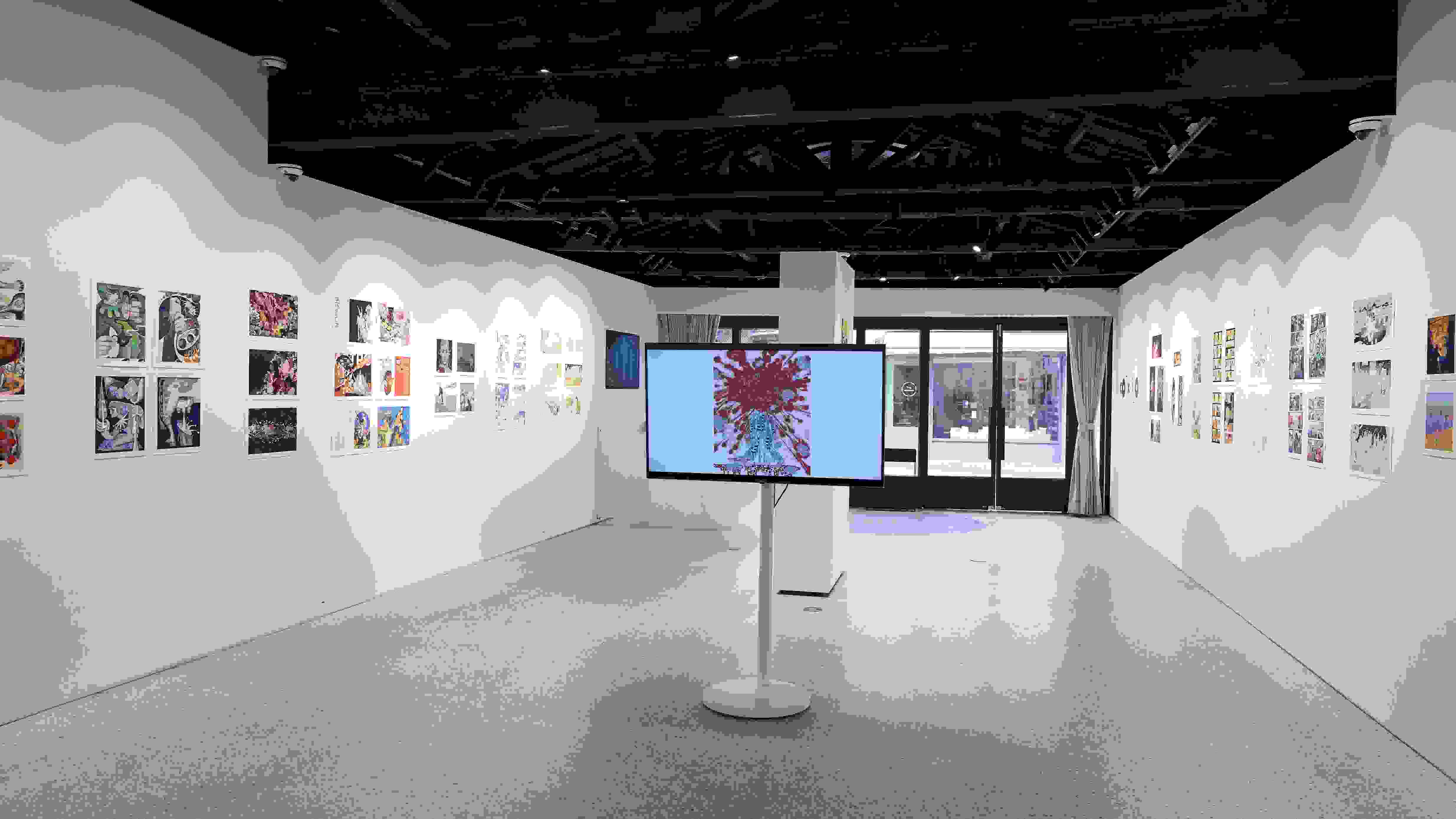 Gallery installation displaying colorful comic and illustration-style artworks arranged linearly along both walls. A large monitor on a white pedestal at the center shows a figure with red abstract patterns. The space features an open ceiling with exposed dark wooden beams, track lighting, and natural light filtering through glass doors at the far end.
