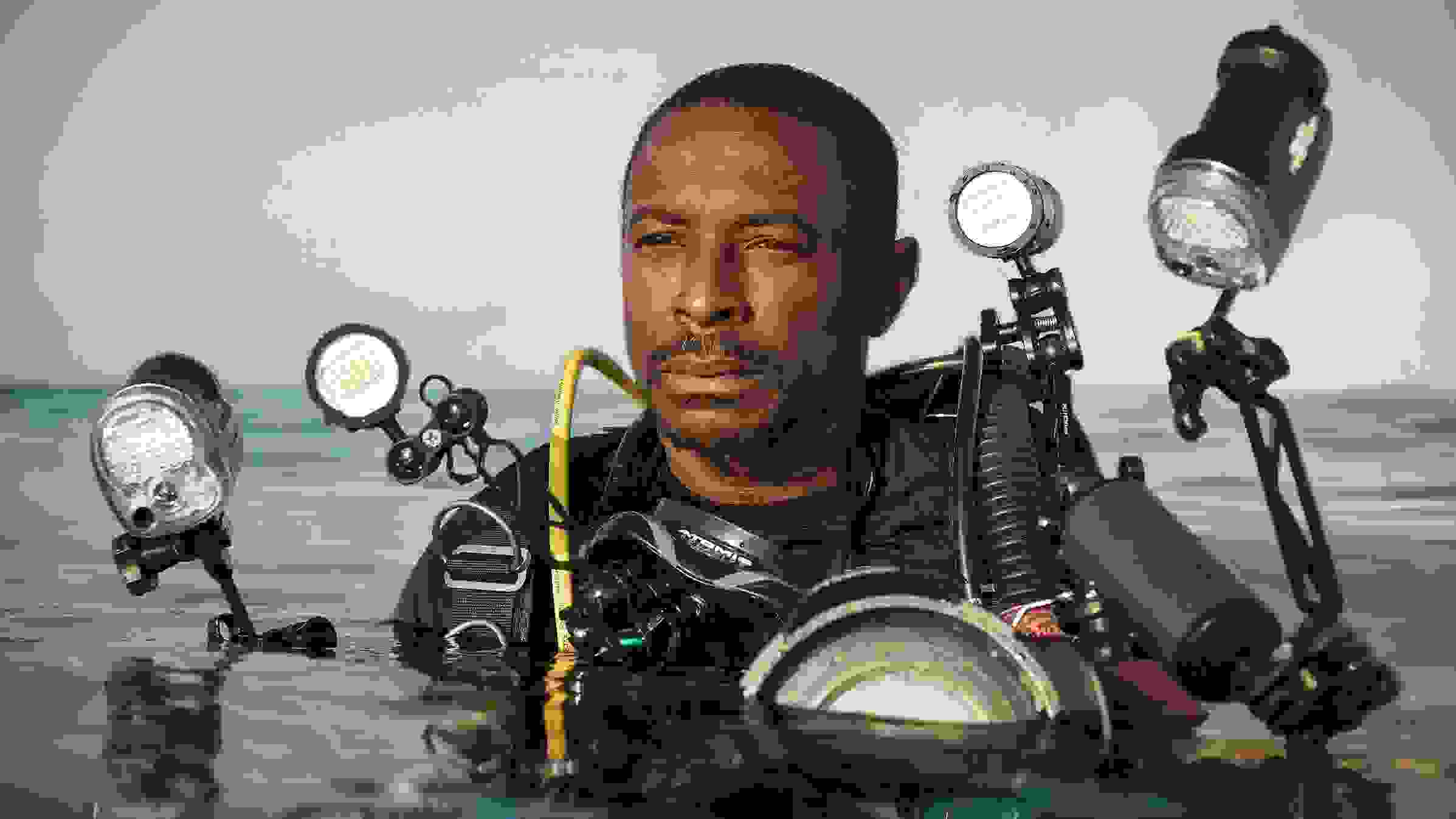 A portrait of a scuba diver in water. He has darker skin and short hair, is wearing a black top, and is equipped with numerous lighting and camera equipments strapped to his body. His body is submerged in water from the chest down.