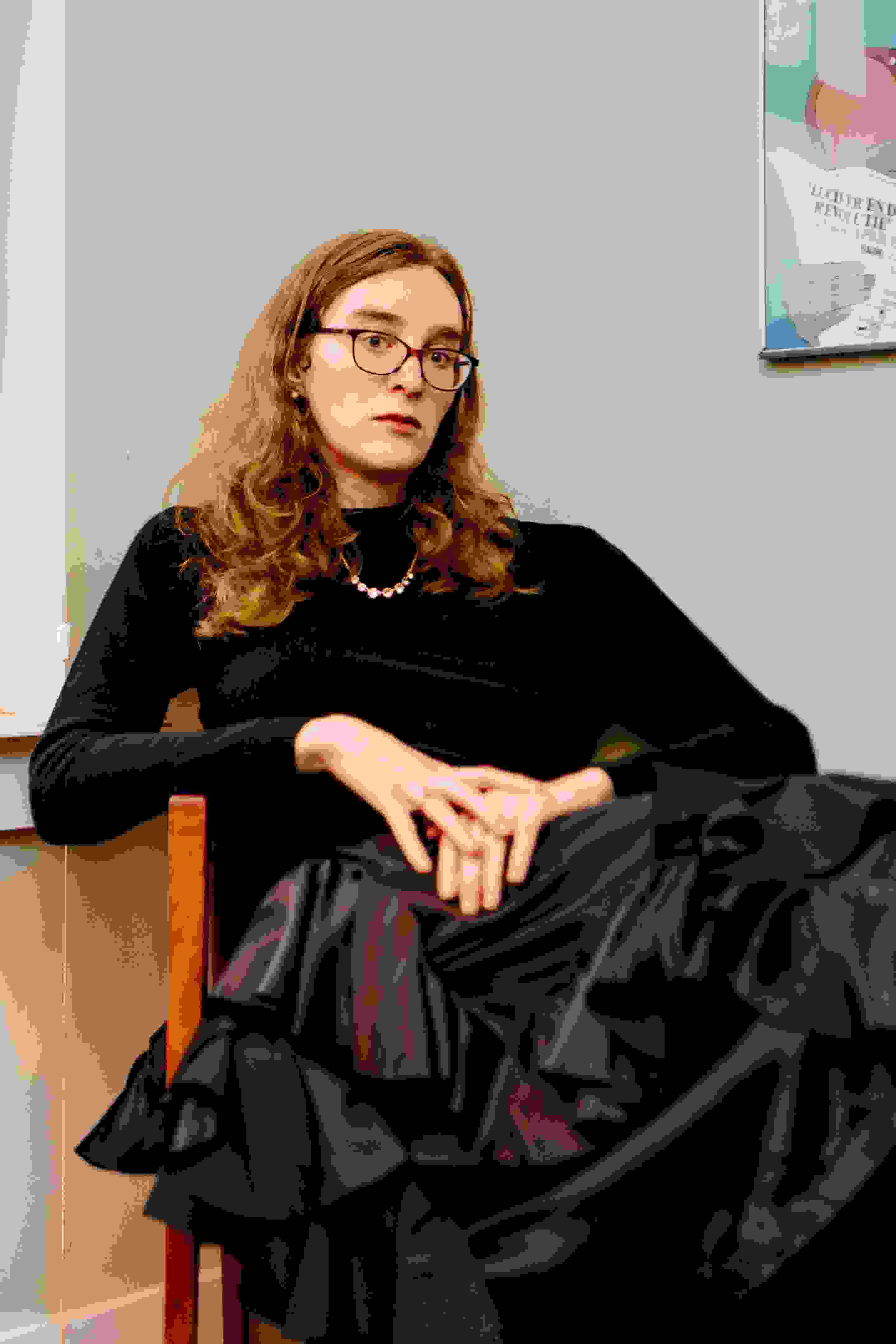 Photo portrait of the writer Grace Byron, sitting in a chair in a black turtleneck with her hands placed on top of one another. Grace wears glasses and looks directly at the lens.