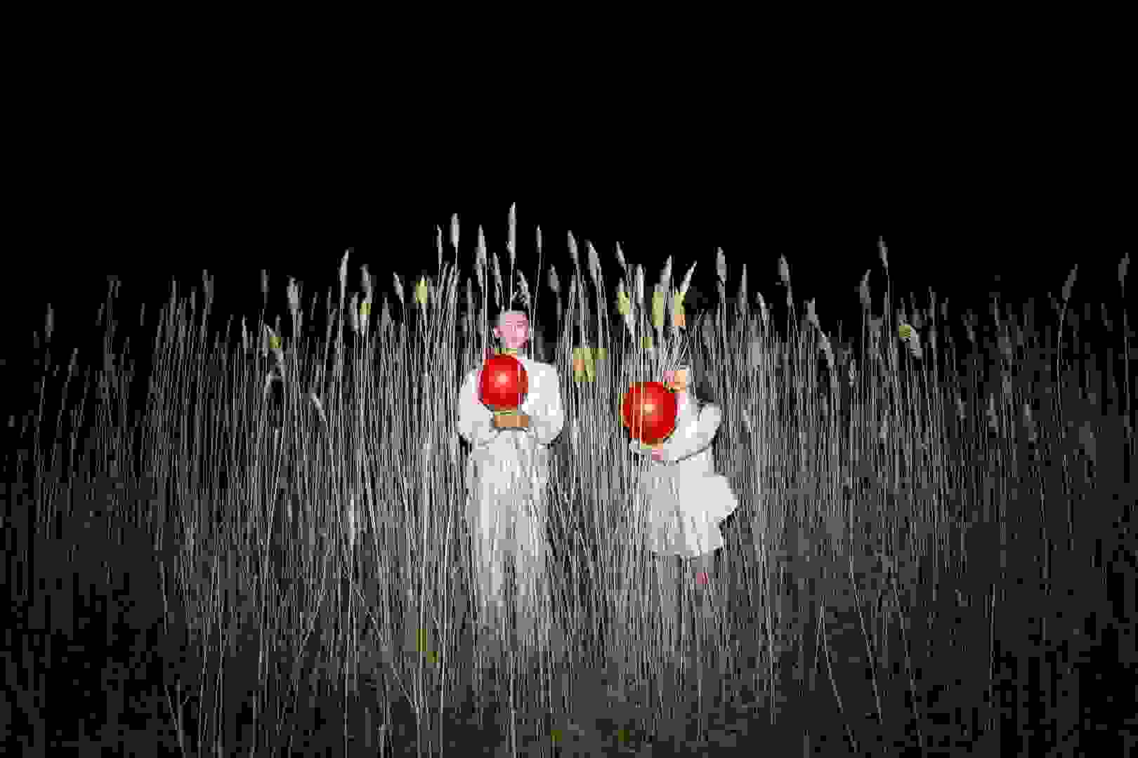 A man and woman wearing all white are each holding a red balloon, standing in tall reeds at night.