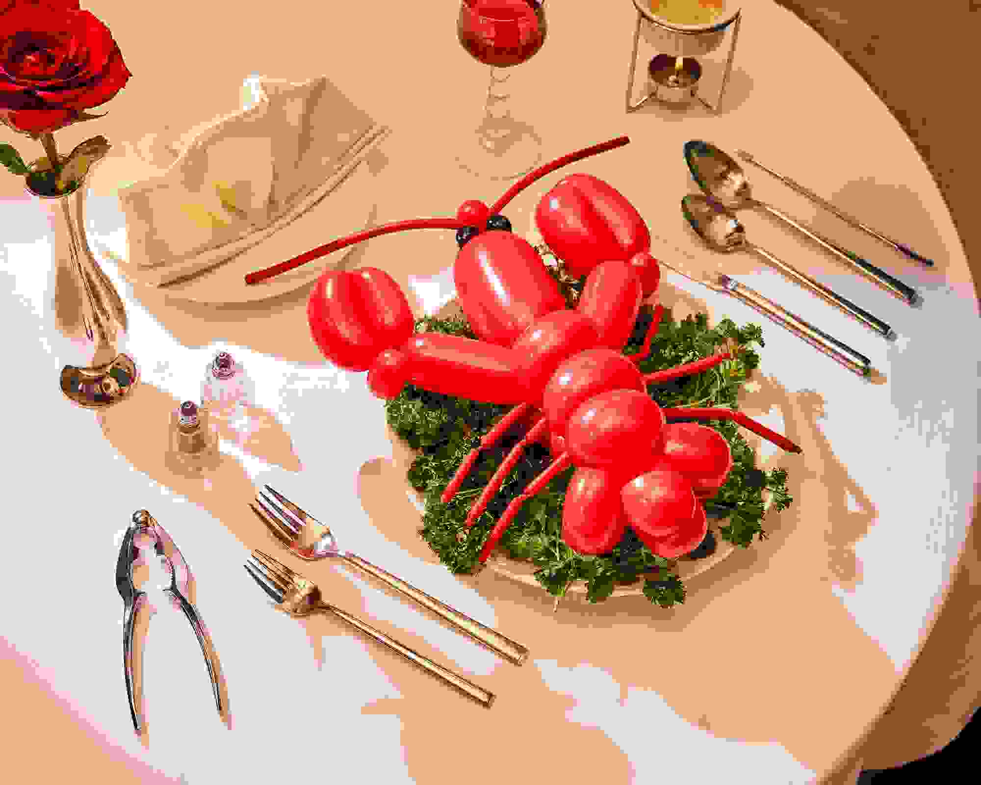 A photograph of a lobster shaped red balloon sitting on top of a bed of kale leaves on a dinner plate. Surrounding the scene is a dinner table fully set up with cutlery,  a dinner napkin folded into a fan, and a single rose in a tin vase.