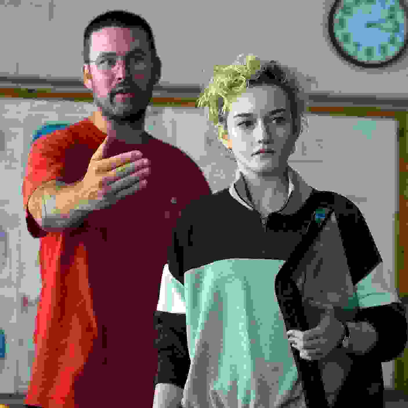 Photograph of a director in a red shirt standing with his hand on his chest beside a teenage actor with short curly blonde hair wearing a navy and gray athletic jacket in a classroom