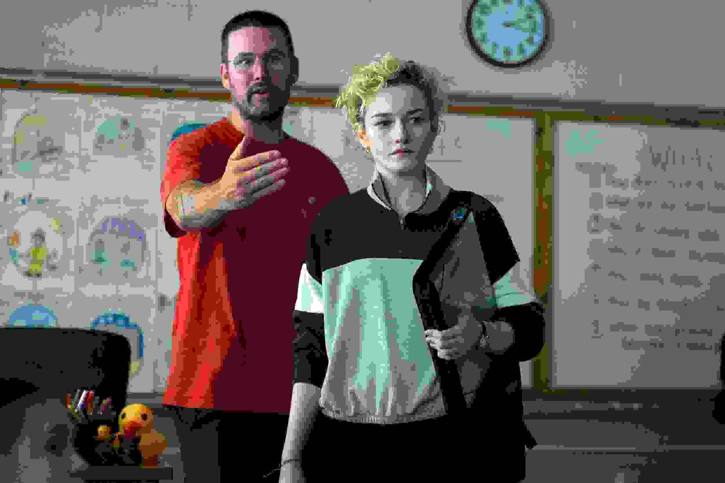 A person in a red shirt gestures in a classroom, appearing explanatory, while another with short curly hair listens intently. A whiteboard and clock are in the background.