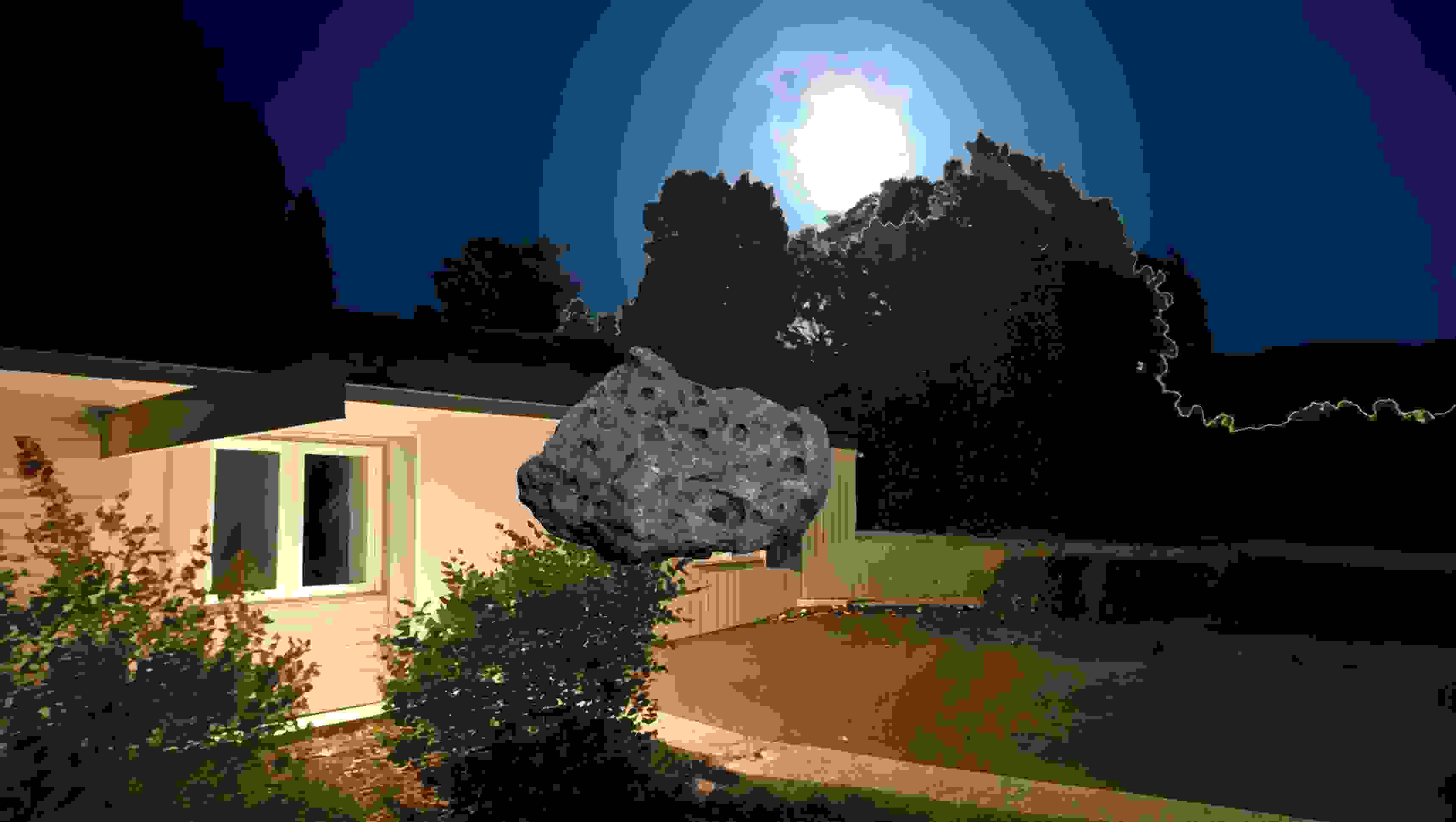 A nighttime scene with a house illuminated by exterior lights. The sky is dark blue with a glowing full moon shining above trees. In the foreground, a large gray rock with crater-like holes appears digitally inserted, floating in mid-air in front of the house.