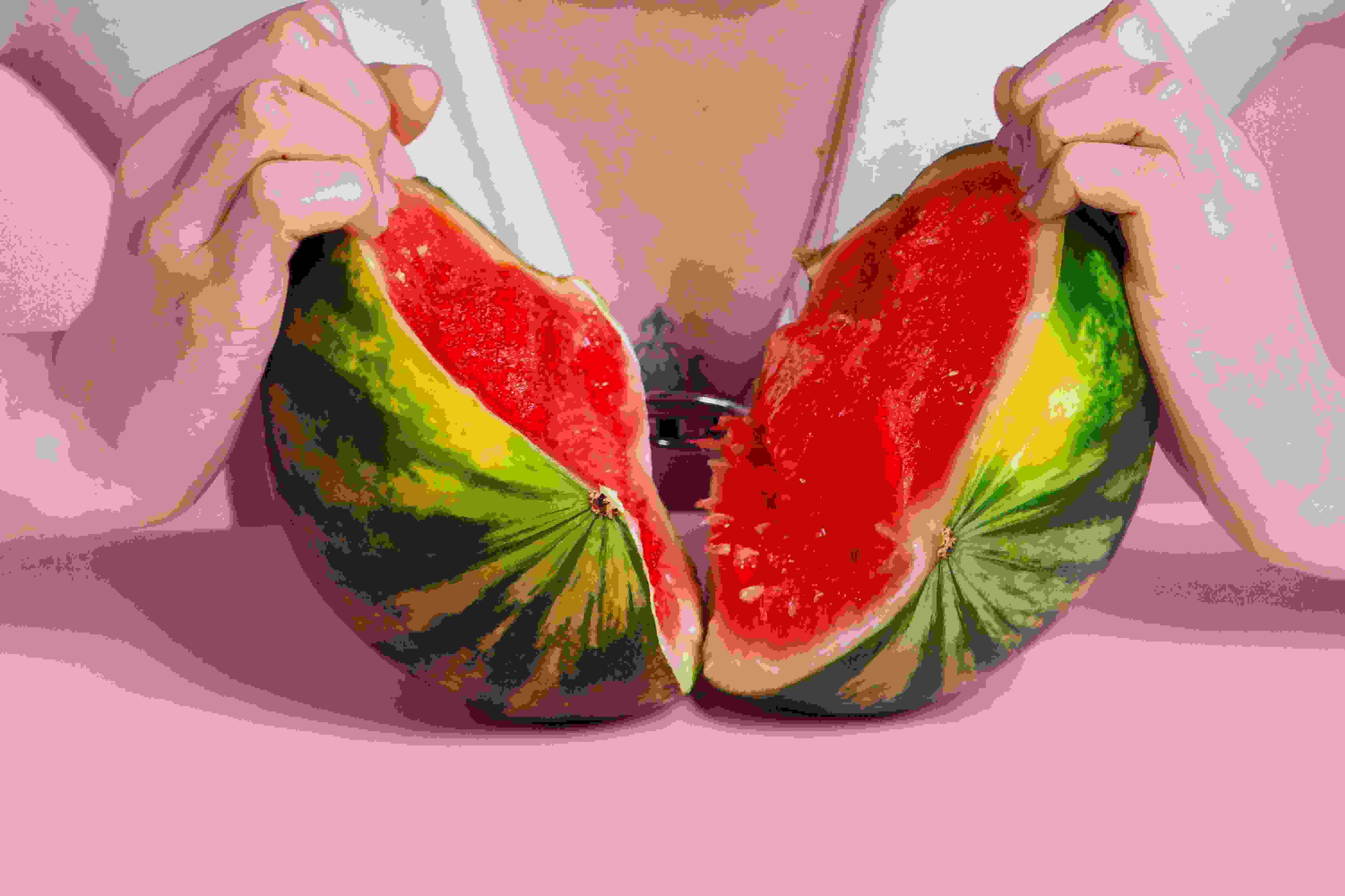 Two hards ripping open a small watermelon in front of a person's chest. There's a pink wall and table in the image as well.