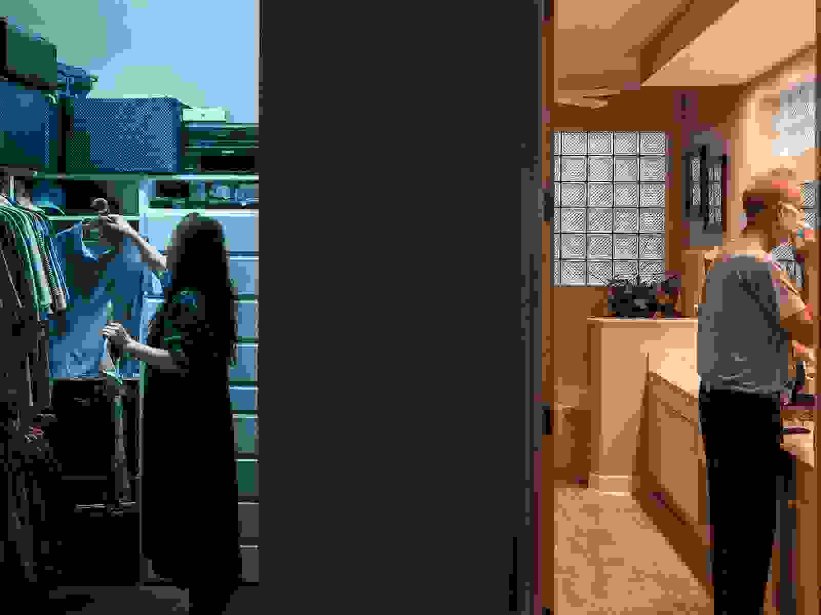 A dark wall in the middle of the image divides this color photograph into three equal parts. In the left third we see into a closet where an Asian woman is putting away a shirt in cool blue light. In the right third a white man is shaving in a bathroom lit by warm light.