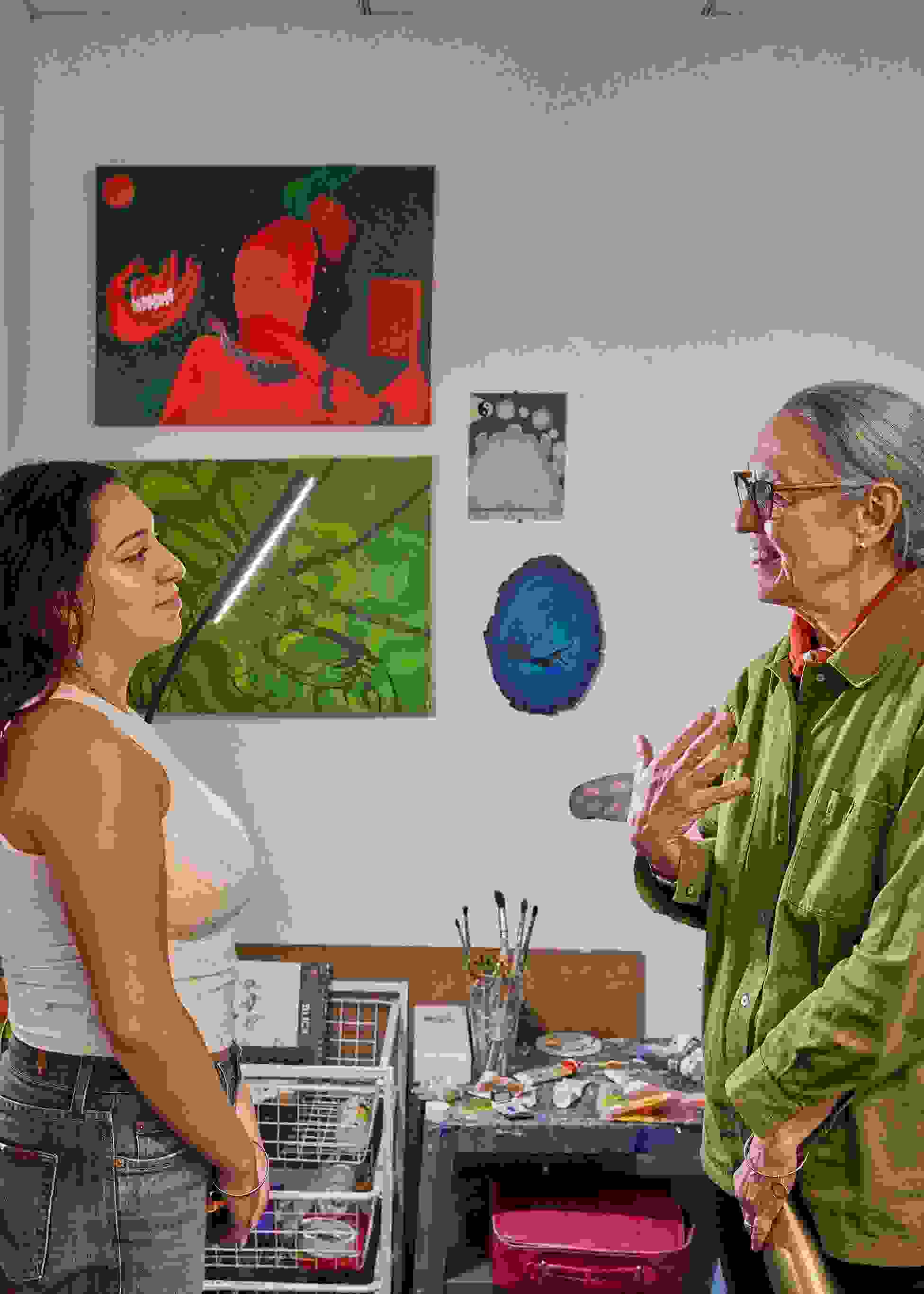 A student and instructor appear to be in conversation; the setting is a painting studio with many canvases, brushes, and other art supplies.
