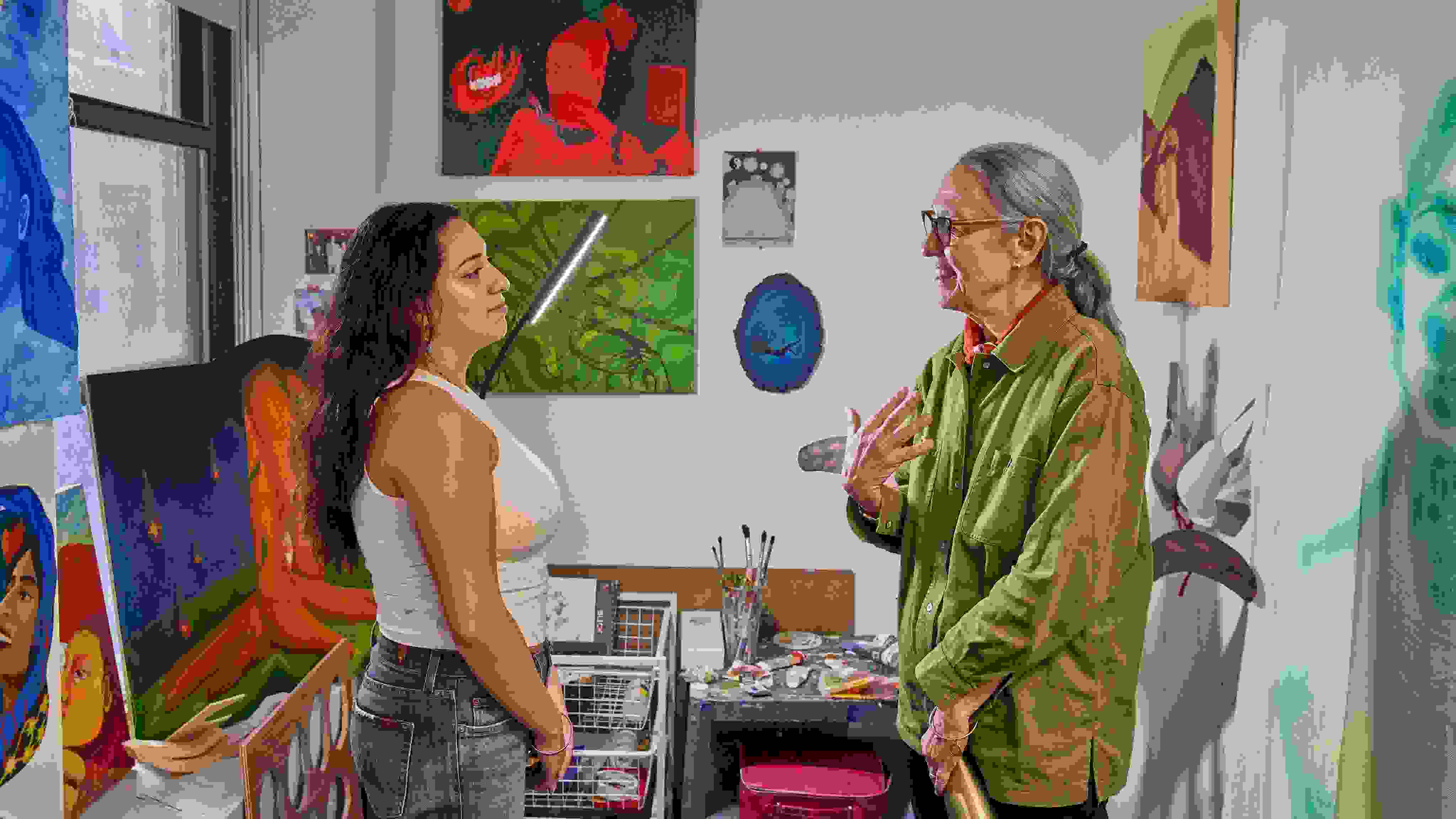 A student and instructor appear to be in conversation; the setting is a painting studio with many canvases, brushes, and other art supplies.