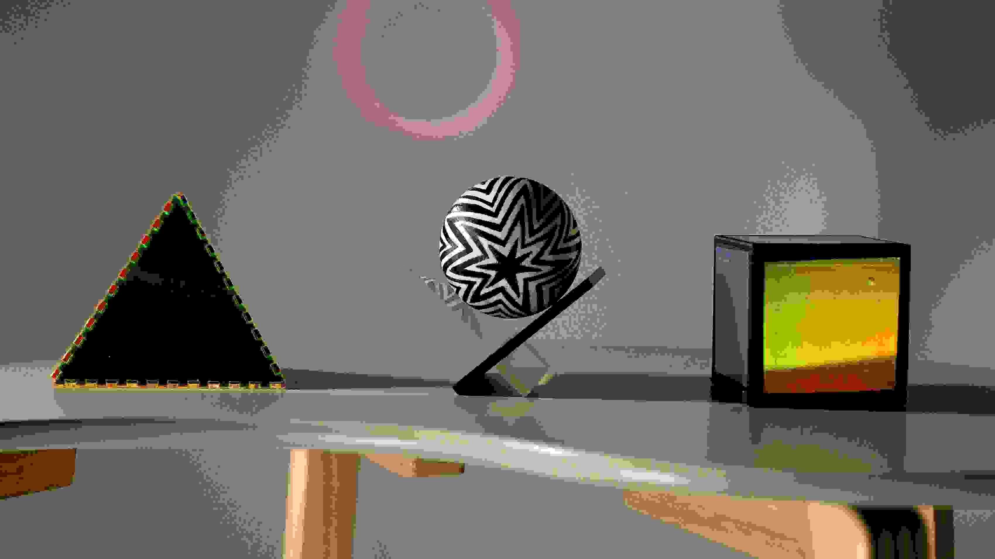 Rendering of three simple objects—a pyramid, a sphere suspended on an acrylic frame, and an iridescent cube—rest on a bean shaped table.