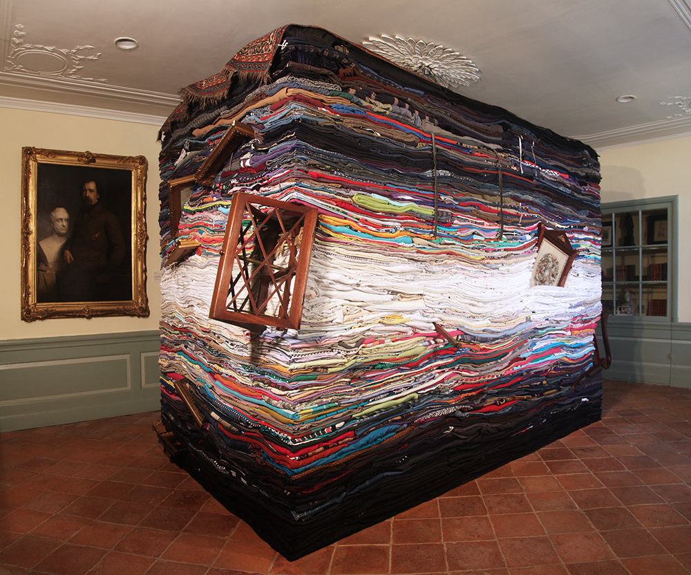 <p "="">A large cuboid sculpture made primarily from stacked and folded secondhand clothes, with wooden furniture emerging from various spots.