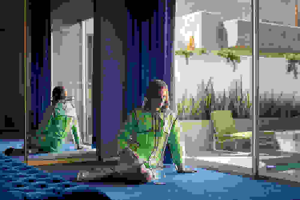 <p "="">A photograph of a person in a green dress and clear plastic raincoat, sitting on the floor next to a glass door looking out onto a swimming pool.