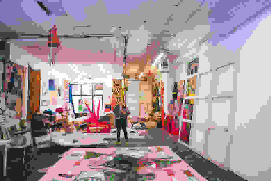 A woman stands in the middle of an art studio filled with large paintings, hanging and stacked rugs, sculptures, pinatas and other objects.