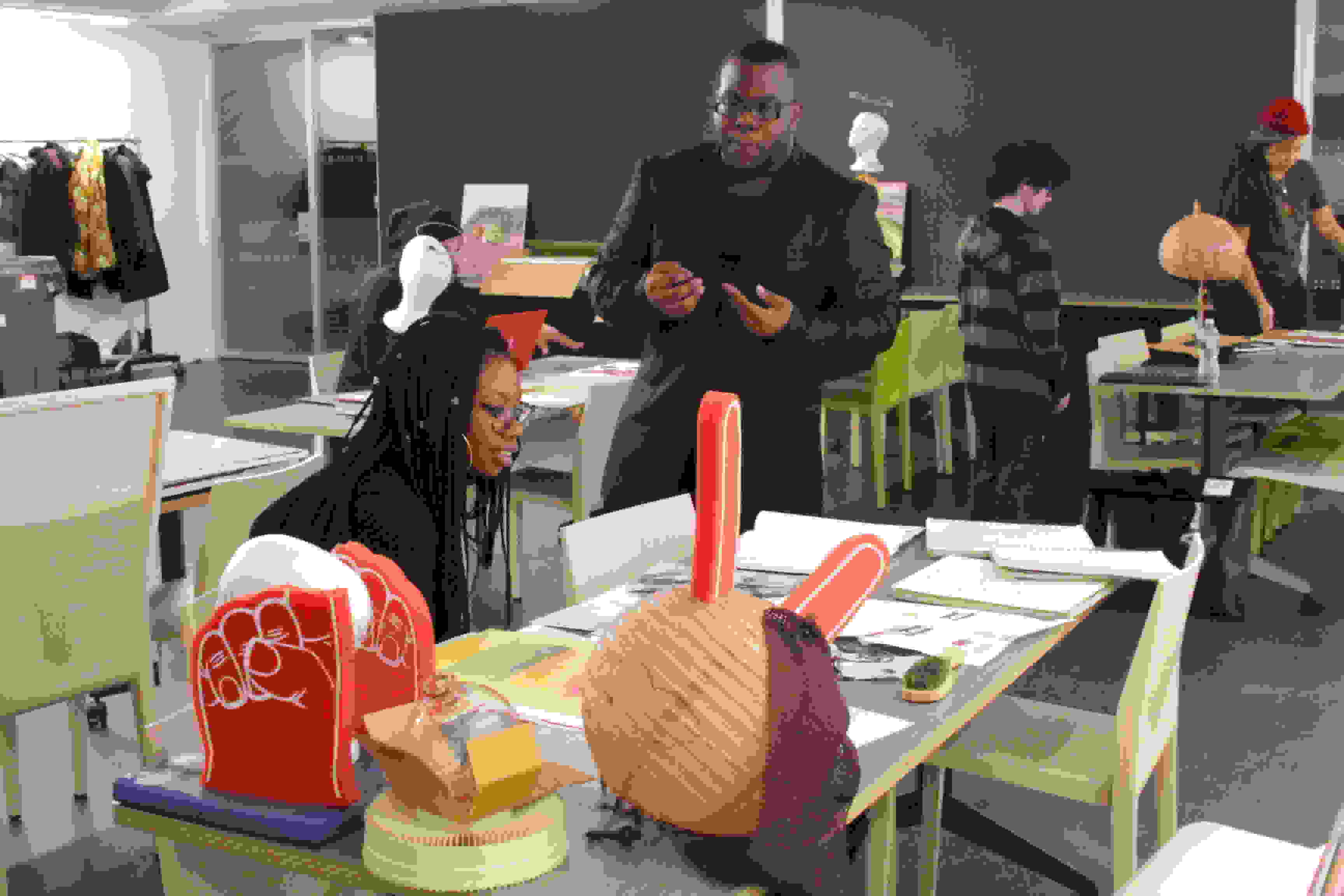 <p "="">A color photograph of several tables with two- and three-dimensional artworks on them. In the foreground, one person sits and looks at the art while another person is standing and gesturing.