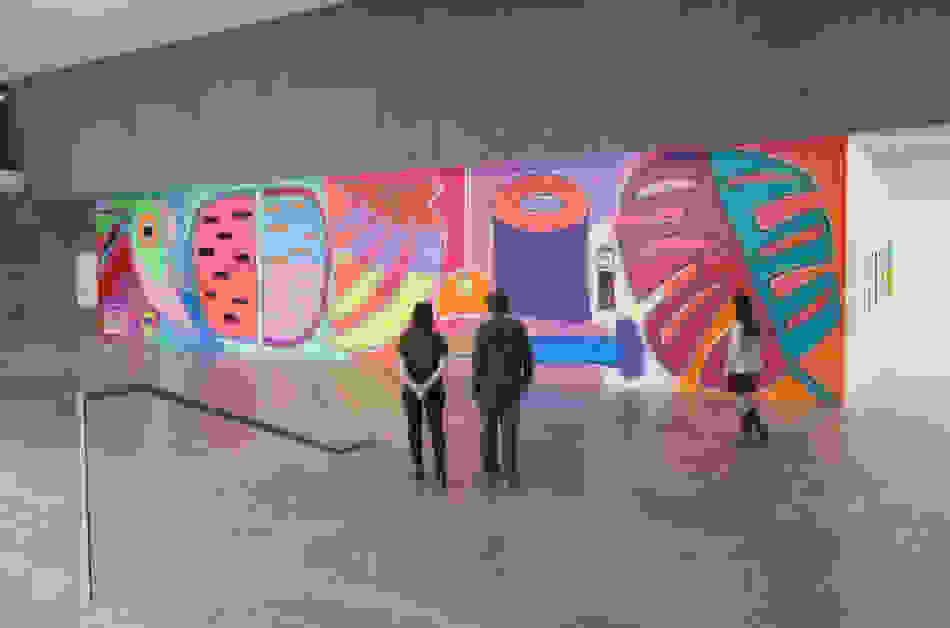 Three museum visitors are seen from behind as they look at a large, colorful mural featuring a tropical bird, watermelon slices, bananas and other objects.