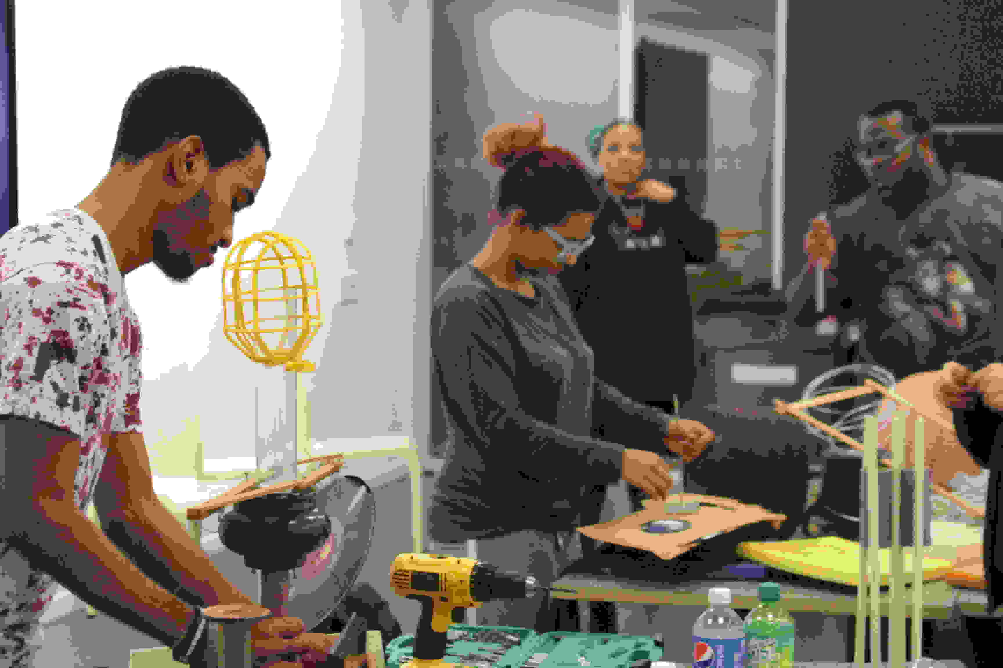 <p "="">Four students stand at or near tables with a variety of art-making materials and tools on them.