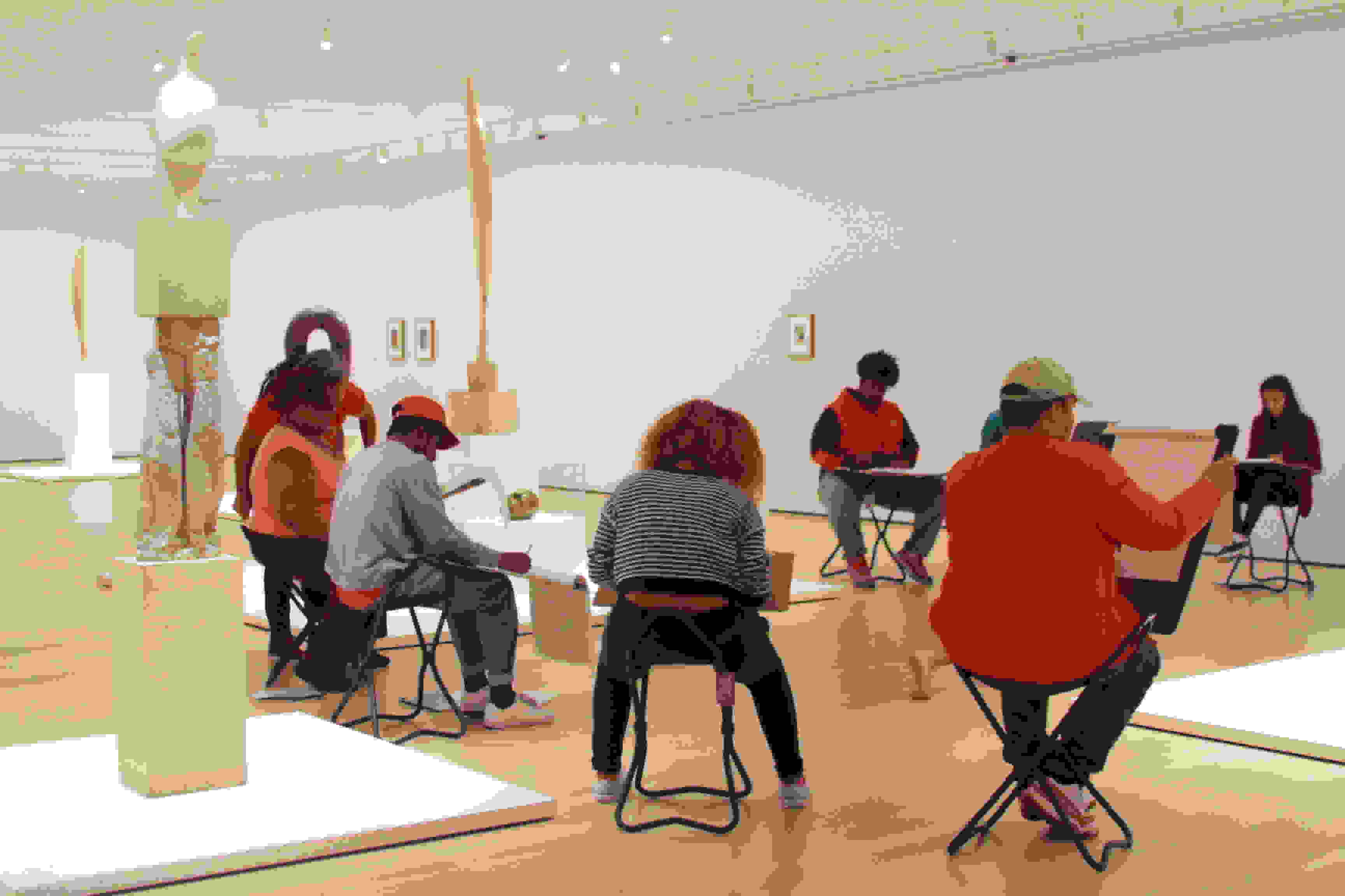 <p "="">A photograph of several art students sitting and sketching amid the sculptures and art on view in a white-walled gallery space.