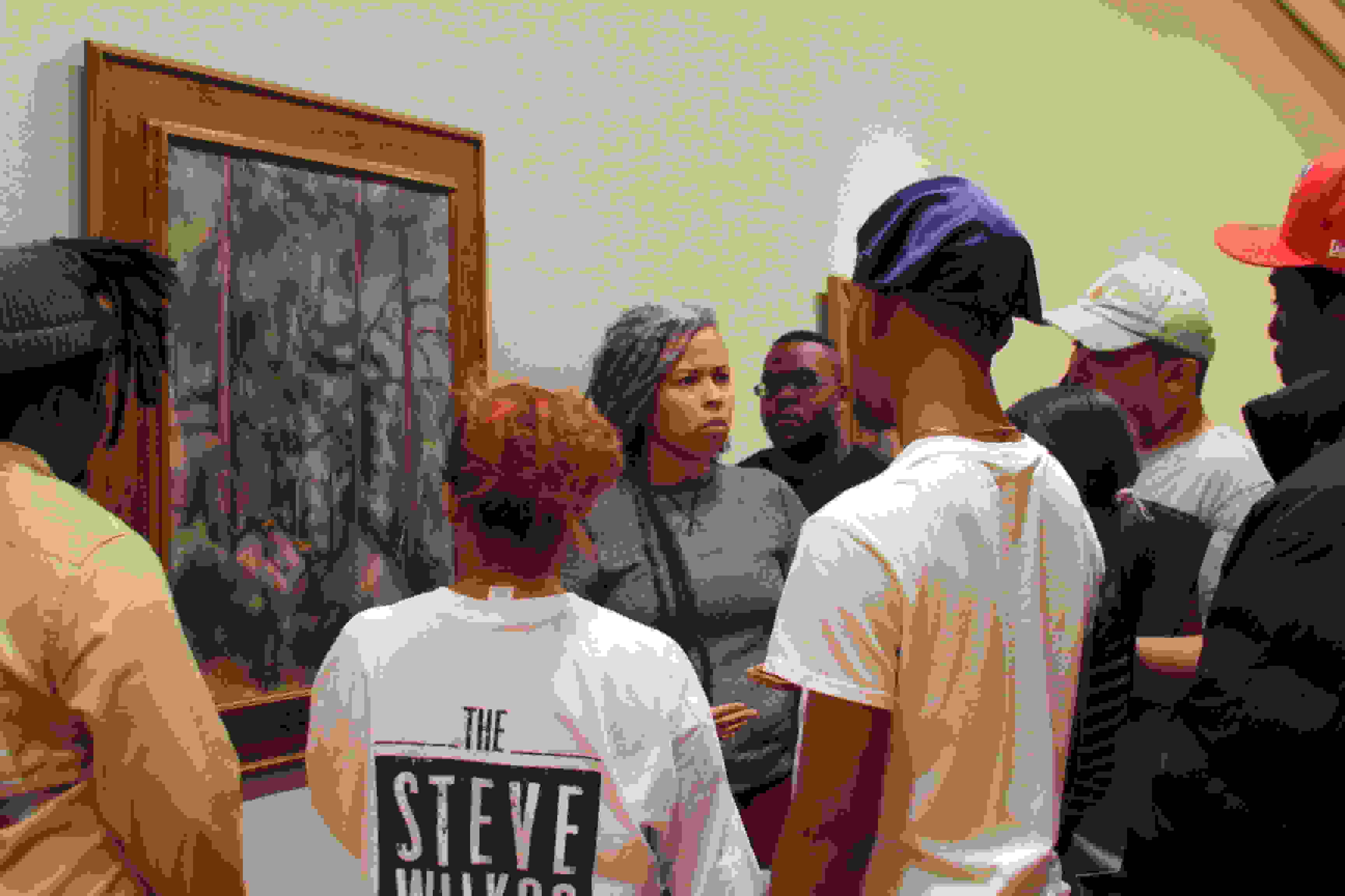 A woman stands next to a painting of a forest scene and talks to a group of people standing around her.