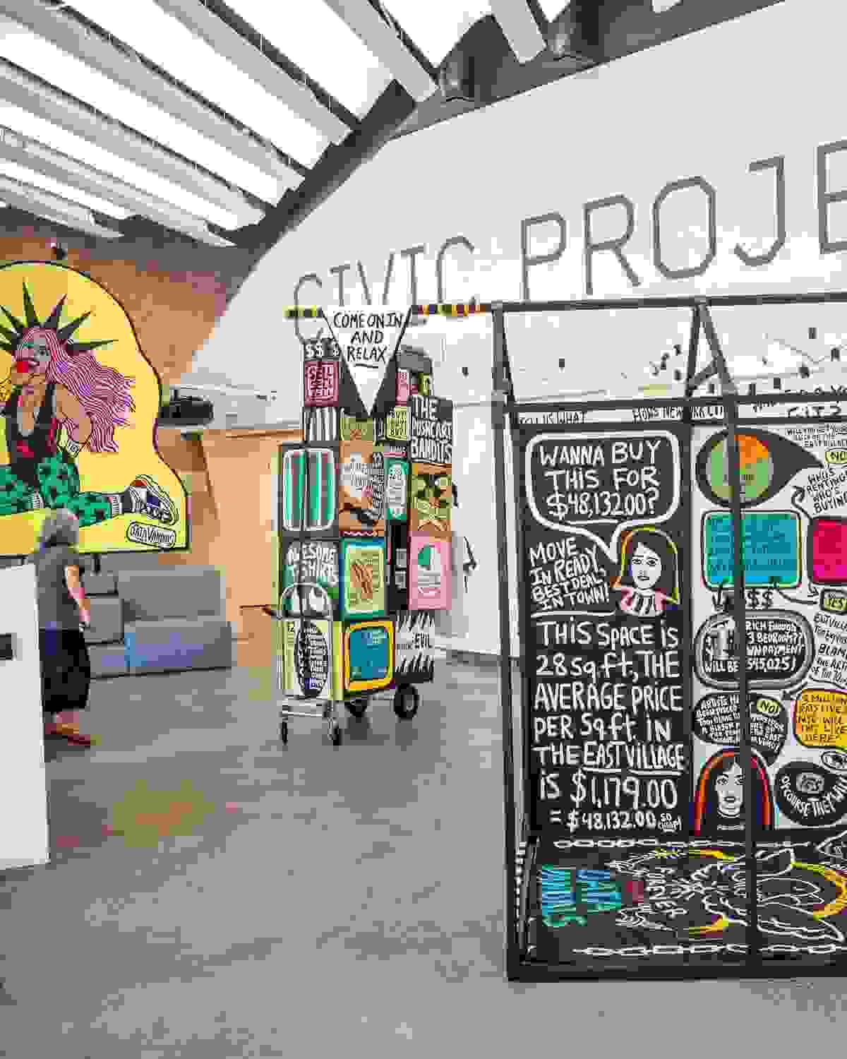 Interactive data visualization installation in a modern gallery space featuring colorful, street art-inspired panels with housing cost data. The central black panel displays NYC housing statistics including '$48,132.00' and text about East Village rental prices per square foot. Surrounding panels feature vibrant graphics with pink, yellow, and green elements. A person in casual clothing examines the display while standing on polished concrete floors under industrial ceiling architecture.