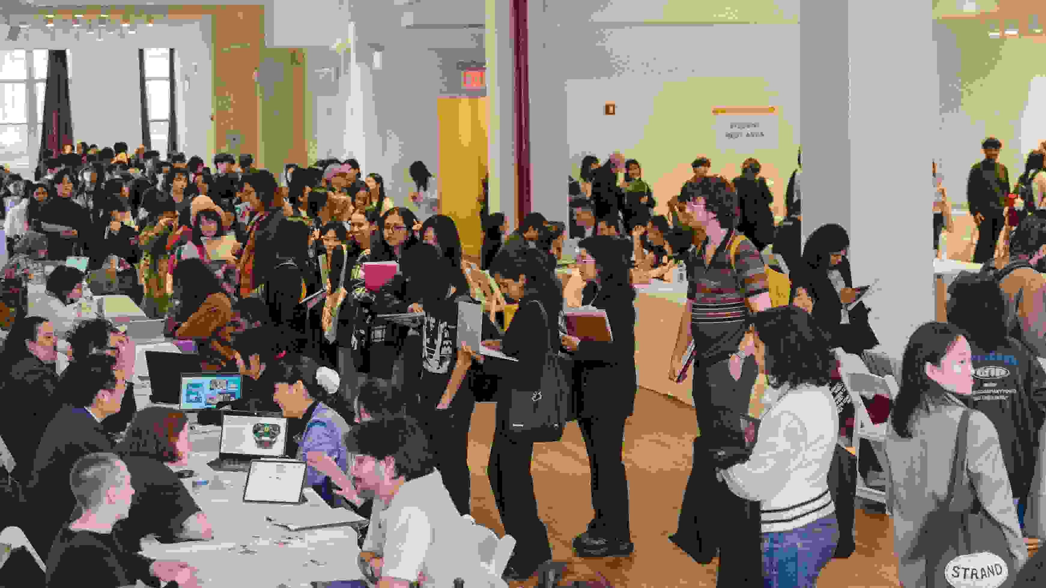 A highly attended career fair event in a large room wherein students mill about from table to table talking to prospective employers