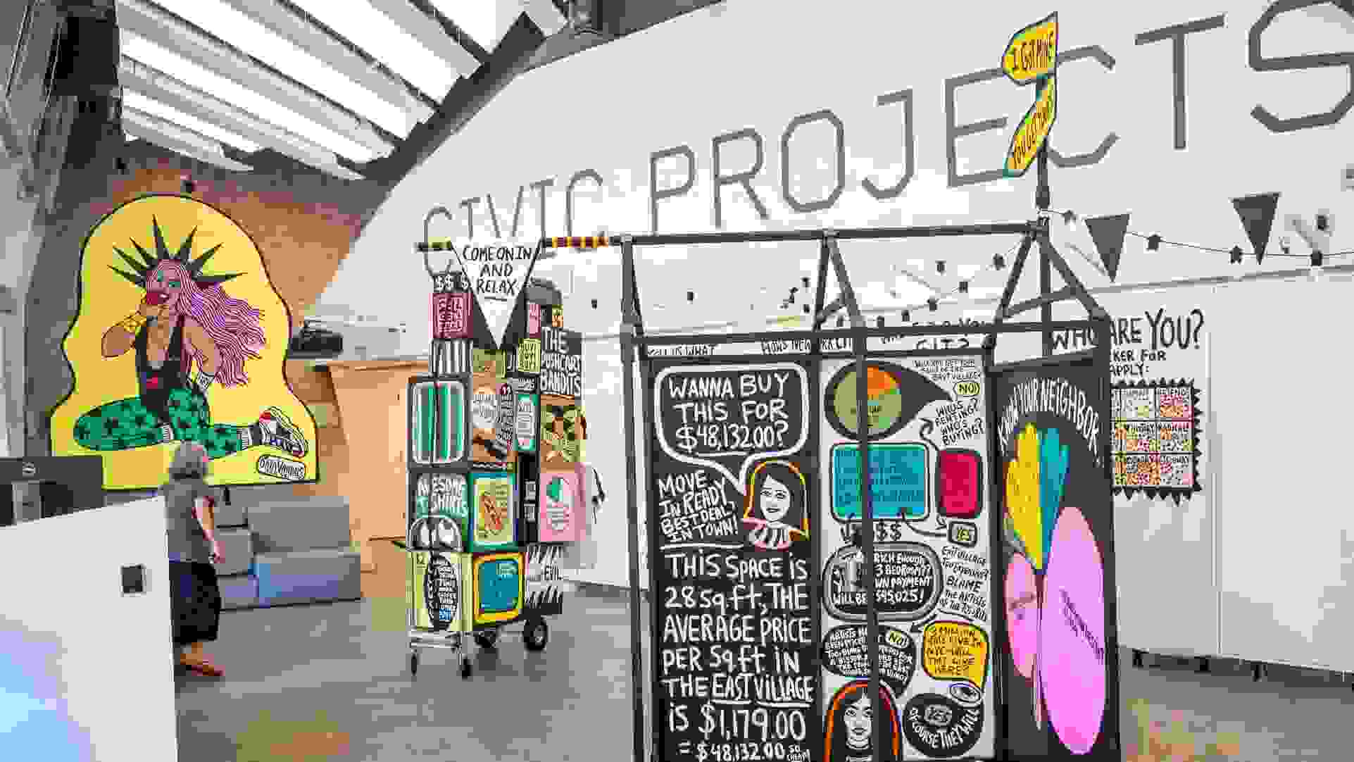 Interactive data visualization installation in a modern gallery space featuring colorful, street art-inspired panels with housing cost data. The central black panel displays NYC housing statistics including '$48,132.00' and text about East Village rental prices per square foot. Surrounding panels feature vibrant graphics with pink, yellow, and green elements. A person in casual clothing examines the display while standing on polished concrete floors under industrial ceiling architecture.