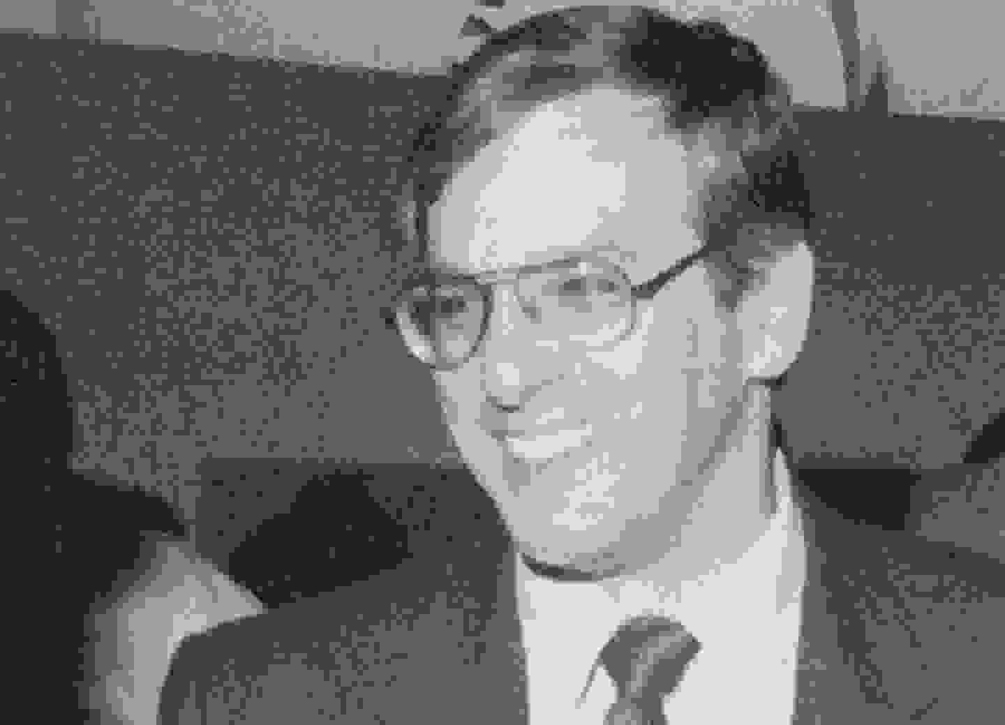 Grainy black and white close up of a man in a suit and tie with aviator prescription glasses.