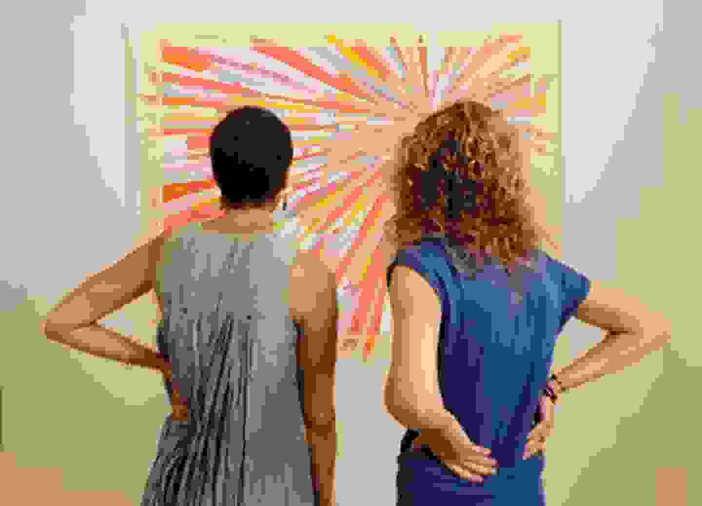 Two people with their backs to the camera look at a piece of art hung on a wall.