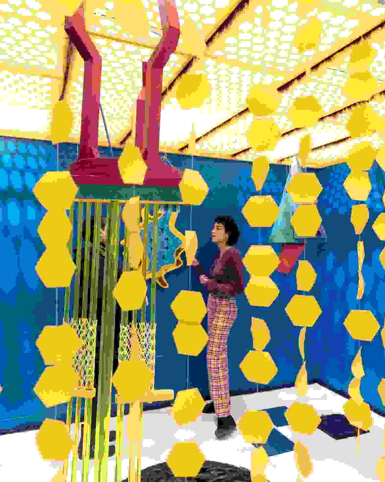 Person standing in an art installation with bright blue walls and mobiles of yellow hexagon-like shapes.