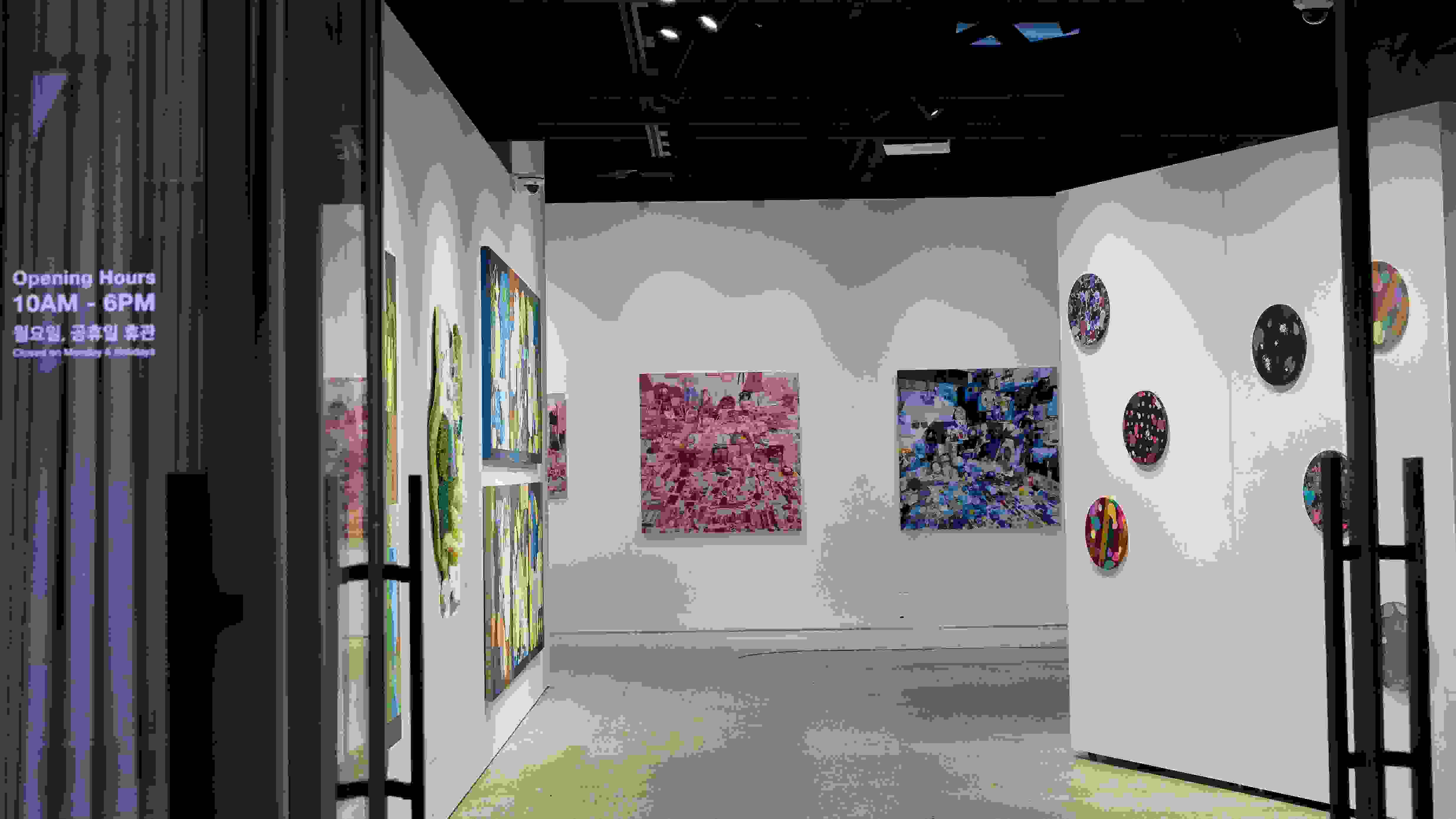 Doors propped open at a gallery's facade, revealing an in-progress exhibition with many art pieces hanging from the three visible walls.