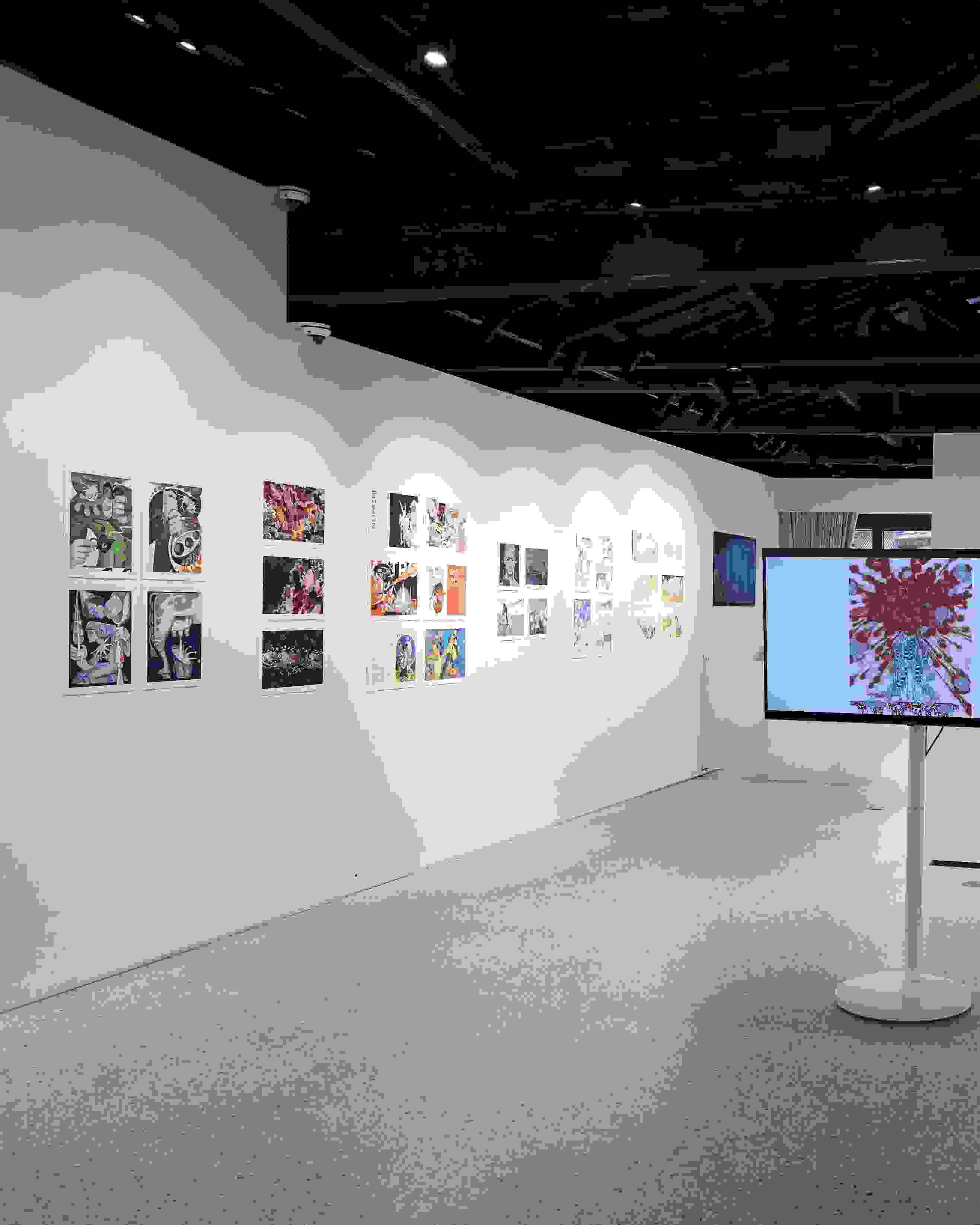 Gallery installation displaying colorful comic and illustration-style artworks arranged linearly along both walls. A large monitor on a white pedestal at the center shows a figure with red abstract patterns. The space features an open ceiling with exposed dark wooden beams, track lighting, and natural light filtering through glass doors at the far end.