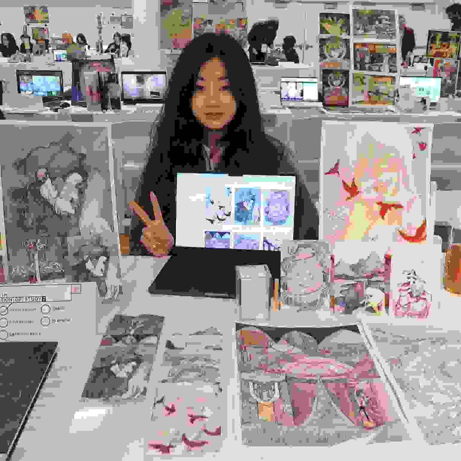A girl poses at a table displaying a variety of artwork.