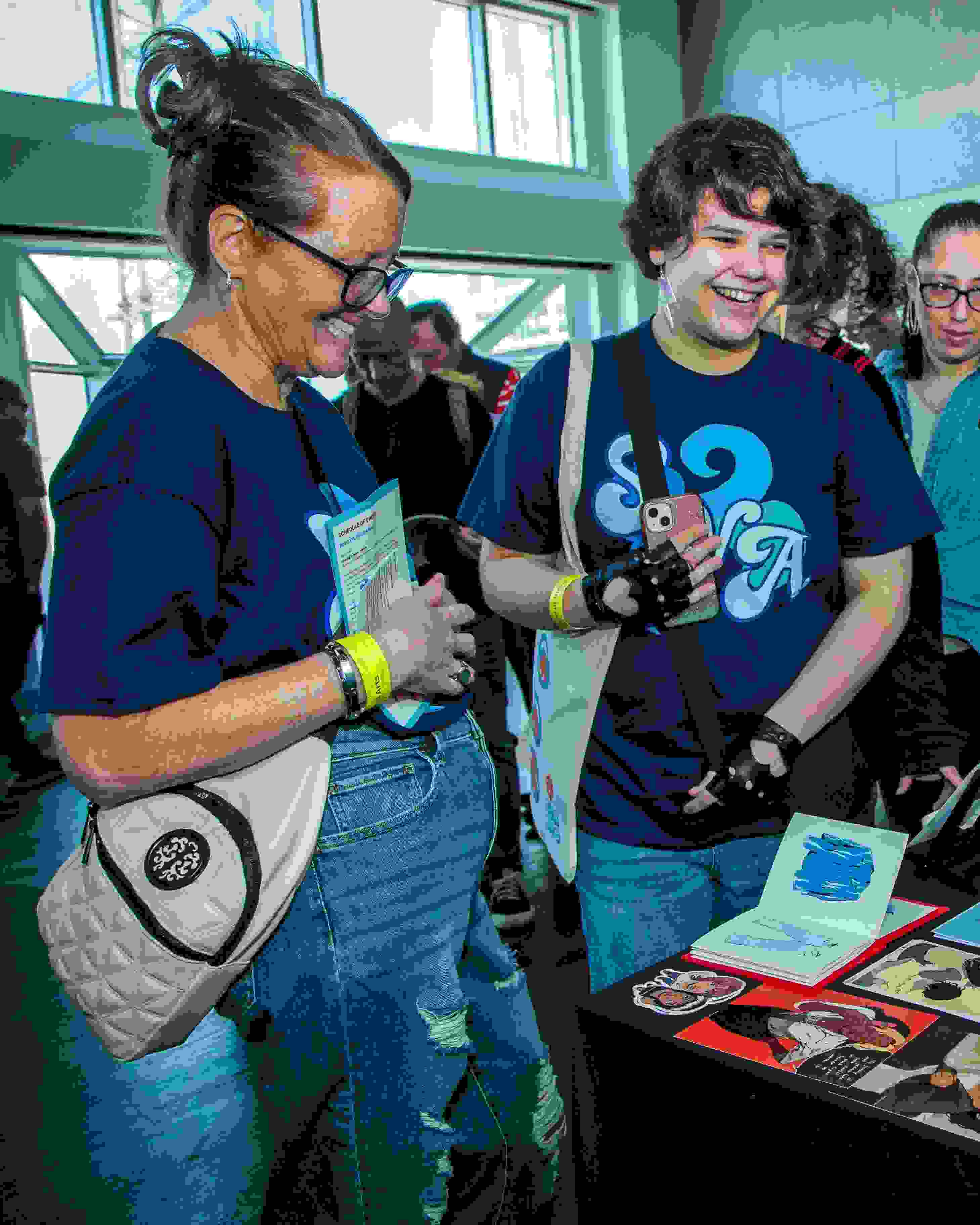 Two people wearing matching shirts are smiling while standing in front of a table of colorful art illustrations