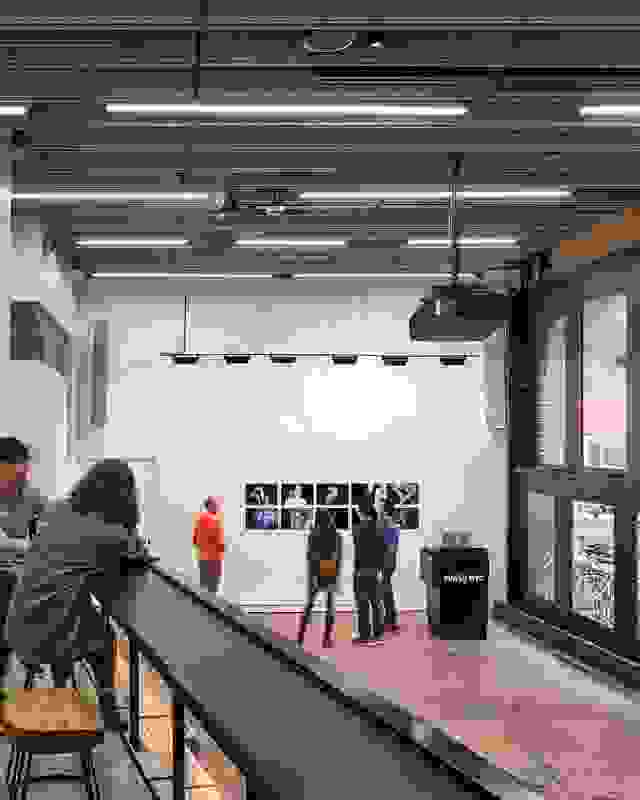 A large room with high ceilings and windows that go from floor to ceiling on the right side of the space. The photo is taken from a mezzanine looking down on the large room. Photographic prints hang on the central wall, with several people gathered around them in the distance. In the foreground, 2 people stand on the mezzanine looking at the same prints. The room has white walls with a dark ceiling and track lighting illuininating the wall of prints. A black podium with a computer atop it sits to the right of th e prints with the SVA NYC logo in blue on the front of it.