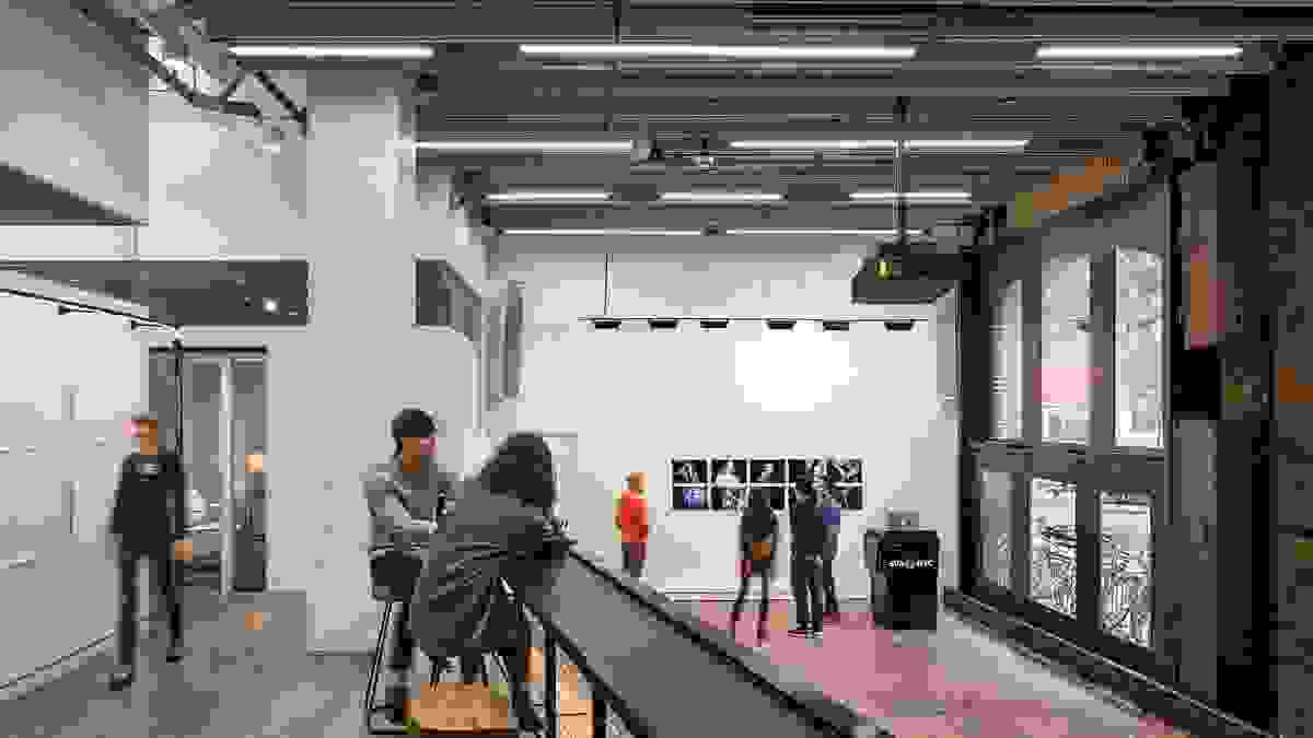 A large room with high ceilings and windows that go from floor to ceiling on the right side of the space. The photo is taken from a mezzanine looking down on the large room. Photographic prints hang on the central wall, with several people gathered around them in the distance. In the foreground, 2 people stand on the mezzanine looking at the same prints. The room has white walls with a dark ceiling and track lighting illuininating the wall of prints. A black podium with a computer atop it sits to the right of th e prints with the SVA NYC logo in blue on the front of it.