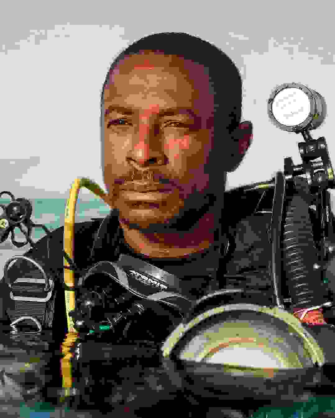 A portrait of a scuba diver in water. He has darker skin and short hair, is wearing a black top, and is equipped with numerous lighting and camera equipments strapped to his body. His body is submerged in water from the chest down.