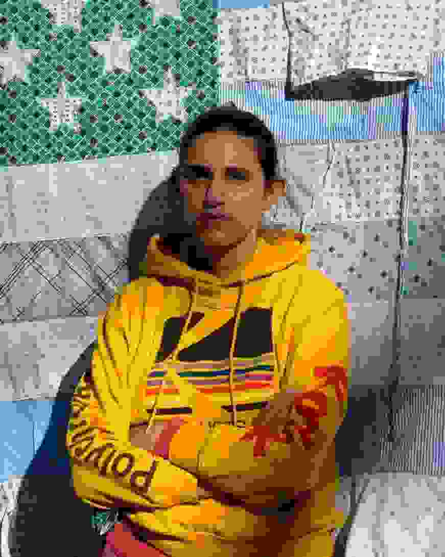 A person in a bright yellow hoodie stands against a quilt resembling an American flag. The quilt has blue and patterned stars and stripes. The mood is calm and contemplative.