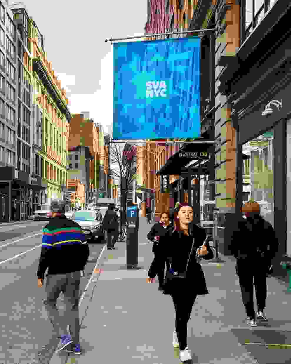 New York City street with pedestrians walking on both sides of the sidewalk as well as crossing the road; two large institutional flags are visible, the left being a red flag, flower outline and the words “SVA NYC,” the left being a blue flag, flower outline and the words “SVA NYC”